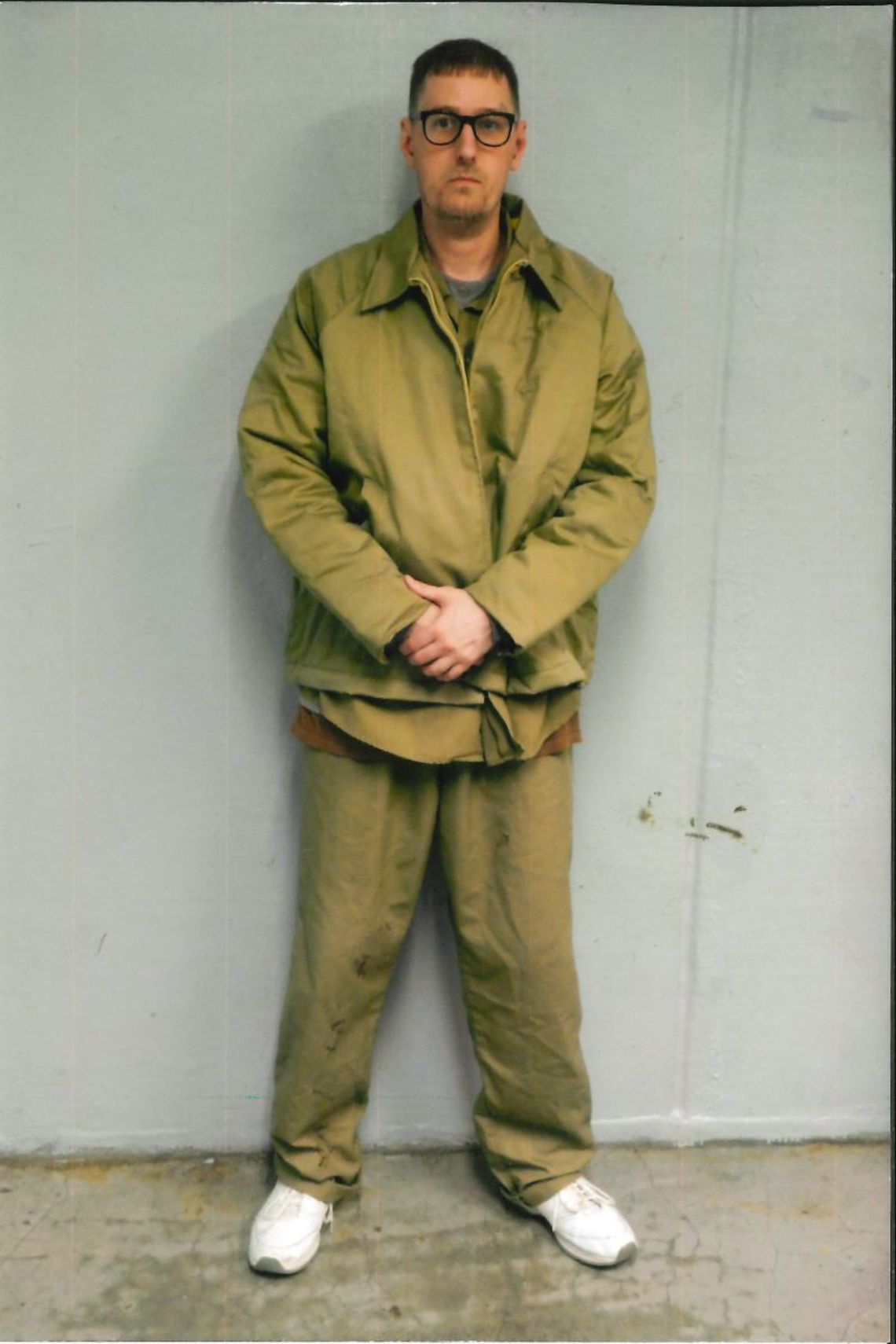 A White man in a green prison suit with white sneakers and black framed glasses stands against a gray wall.