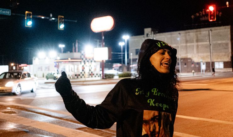 A photo of a Black woman shouting with her fist up while wearing a black hoodie with green text and an image. The woman is standing near an intersection with streetlights at night.