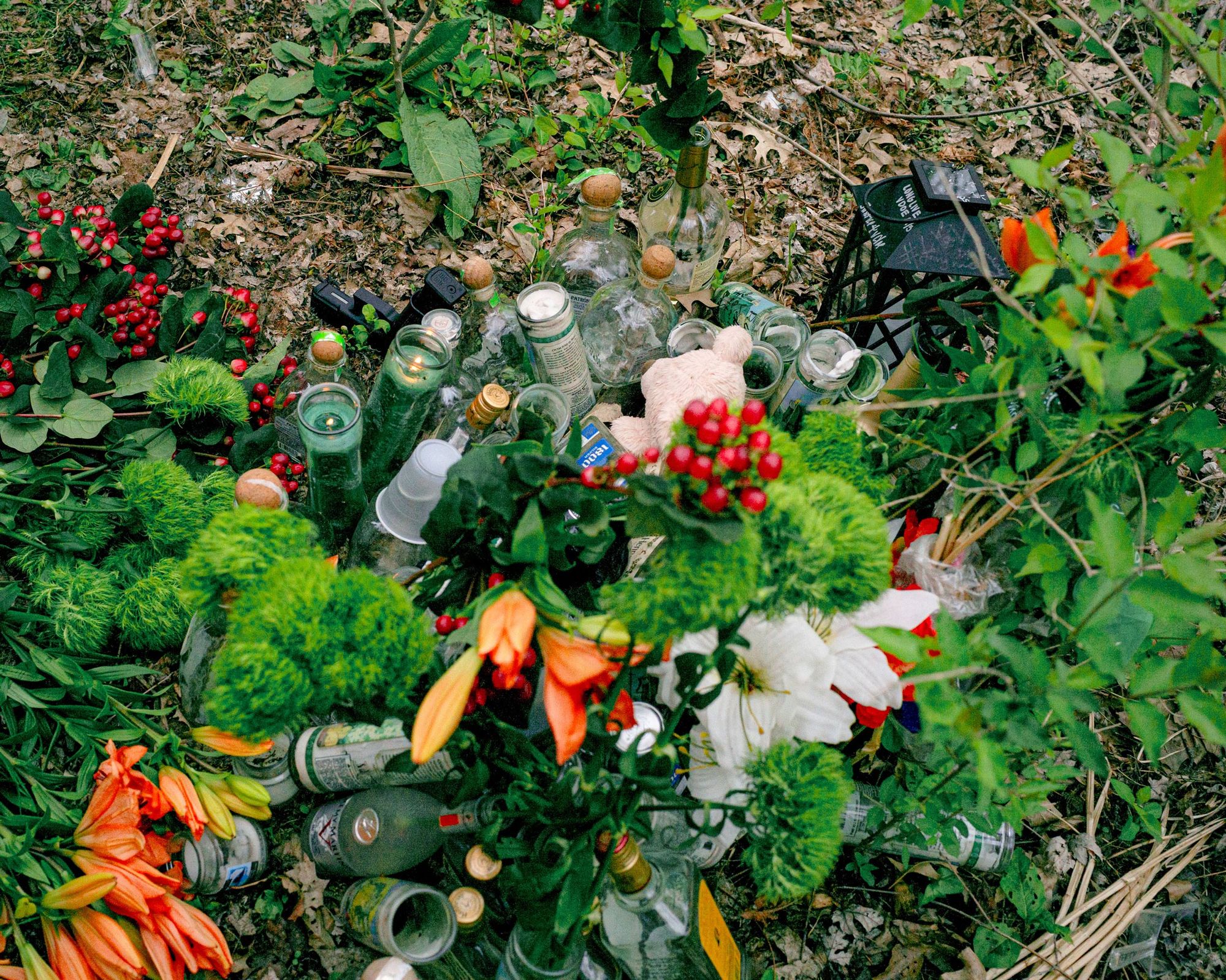 A photo of memorial flowers, candles and drinks on the ground outdoors.