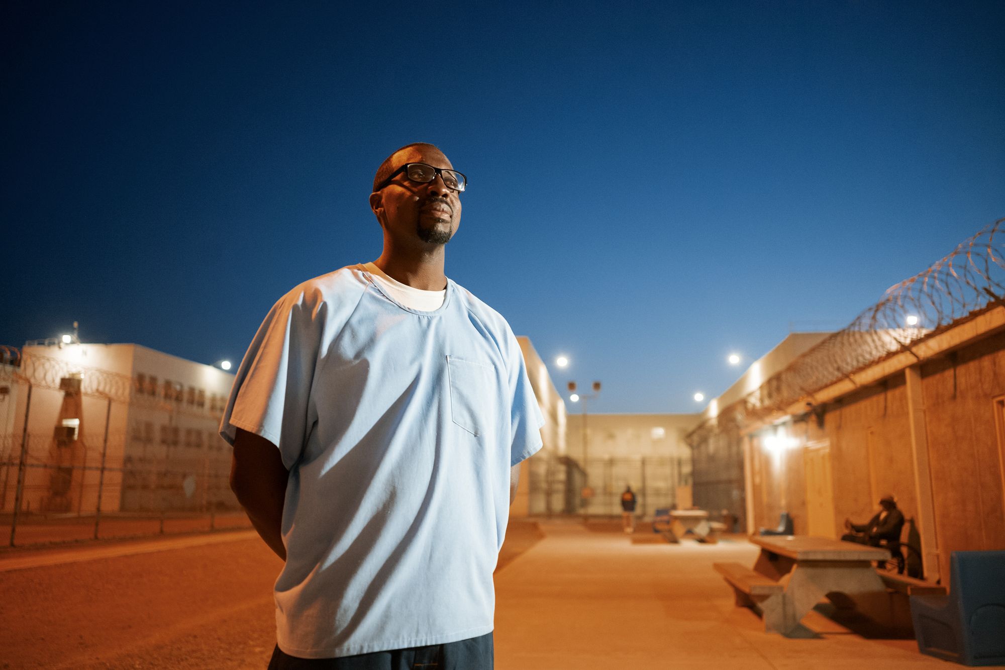 A photo shows a man with dark-toned skin and glasses, wearing a light blue shirt. Barbed wire is visible behind him, in addition to some outdoor tables and people in the background. 