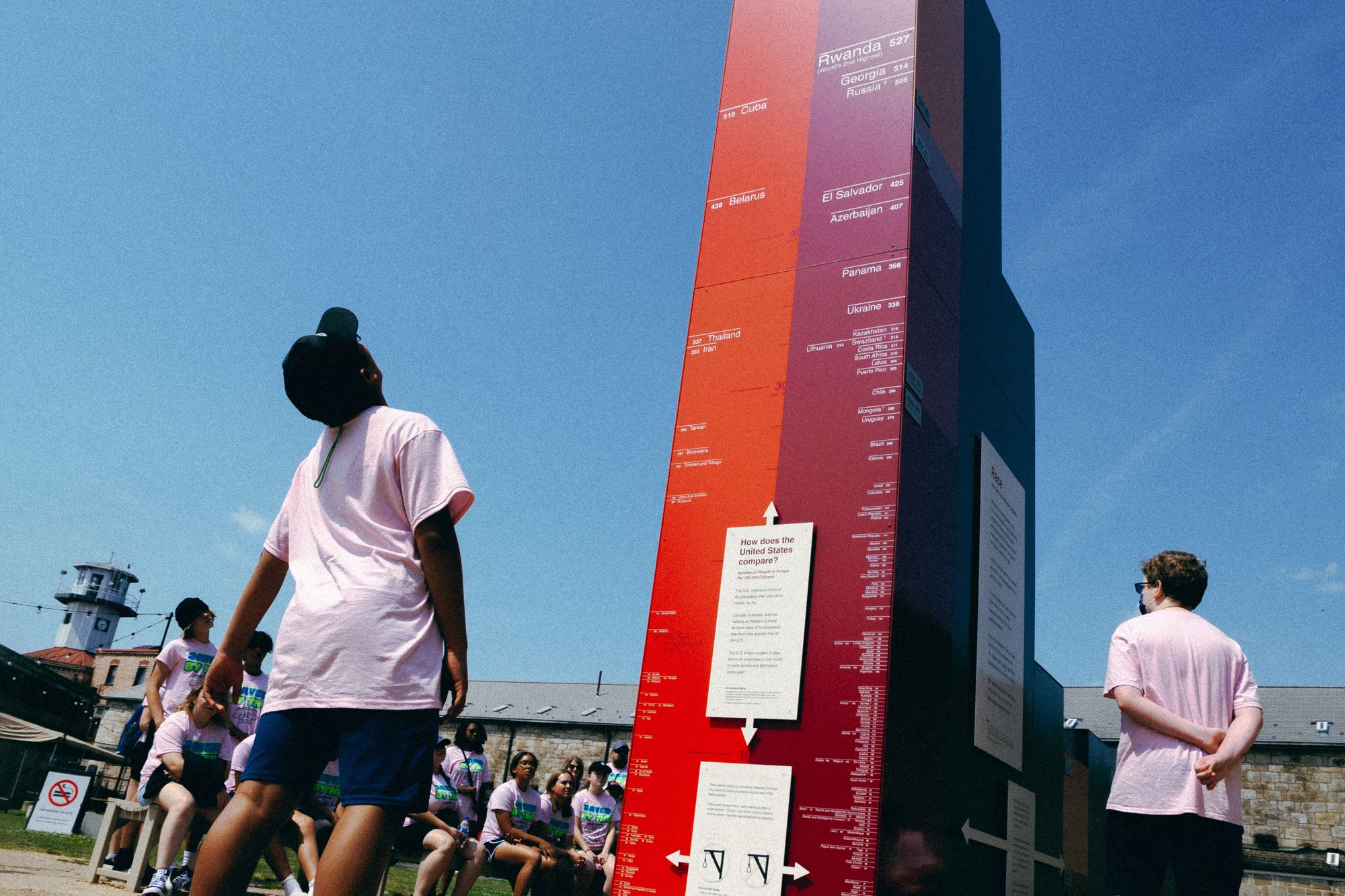 A visitor in a pink shirt, blue shorts, and baseball cap looks up at the tall, red and rectangular sculpture. Other visitors look on. 