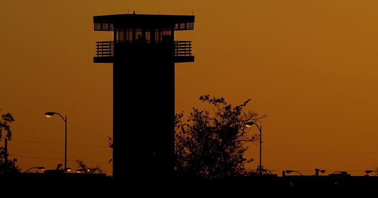 A photo shows the silhouette of a prison tower, trees and lamp posts against an orange sunset. 