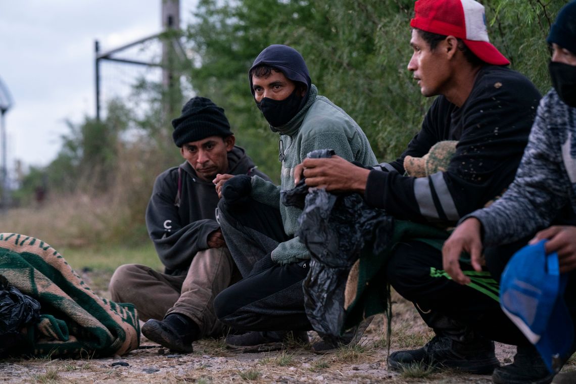 Four people with medium-toned skin sit or crouch on the ground holding clothing and bags. One man is wearing a black cloth mask. 