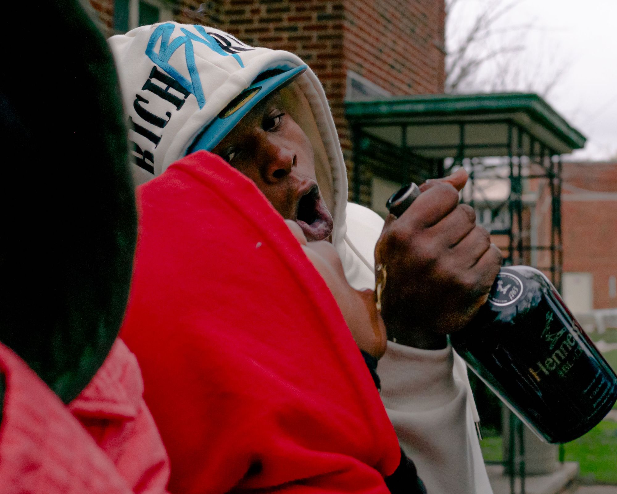 A photo of a young Black man wearing a white hoodie and blue hat, pouring Hennessy into the mouth of another young Black man wearing a red hoodie. 