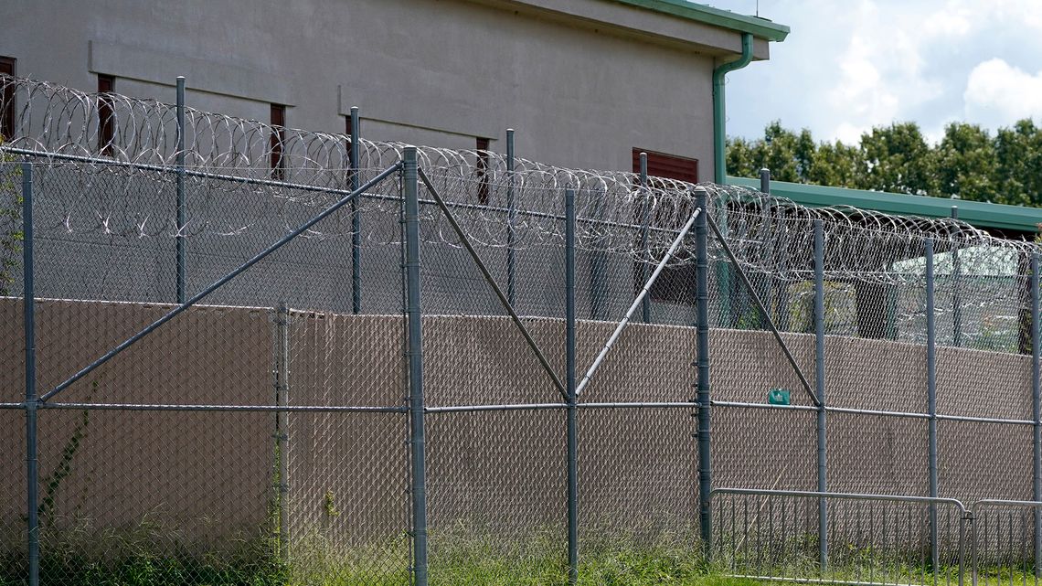 A gray detention center with pale green gutters, behind a barbed wire fence. 