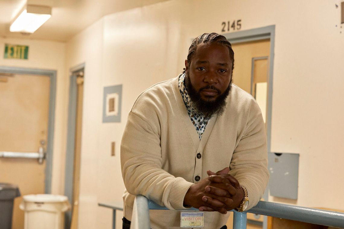 Ofonzo Staton, a Black man with a beard and goatee, wears a cream-colored cardigan and black pants. He leans against a railing painted light blue with a visitor's badge marked "11", looking towards the viewer with a pensive expression.