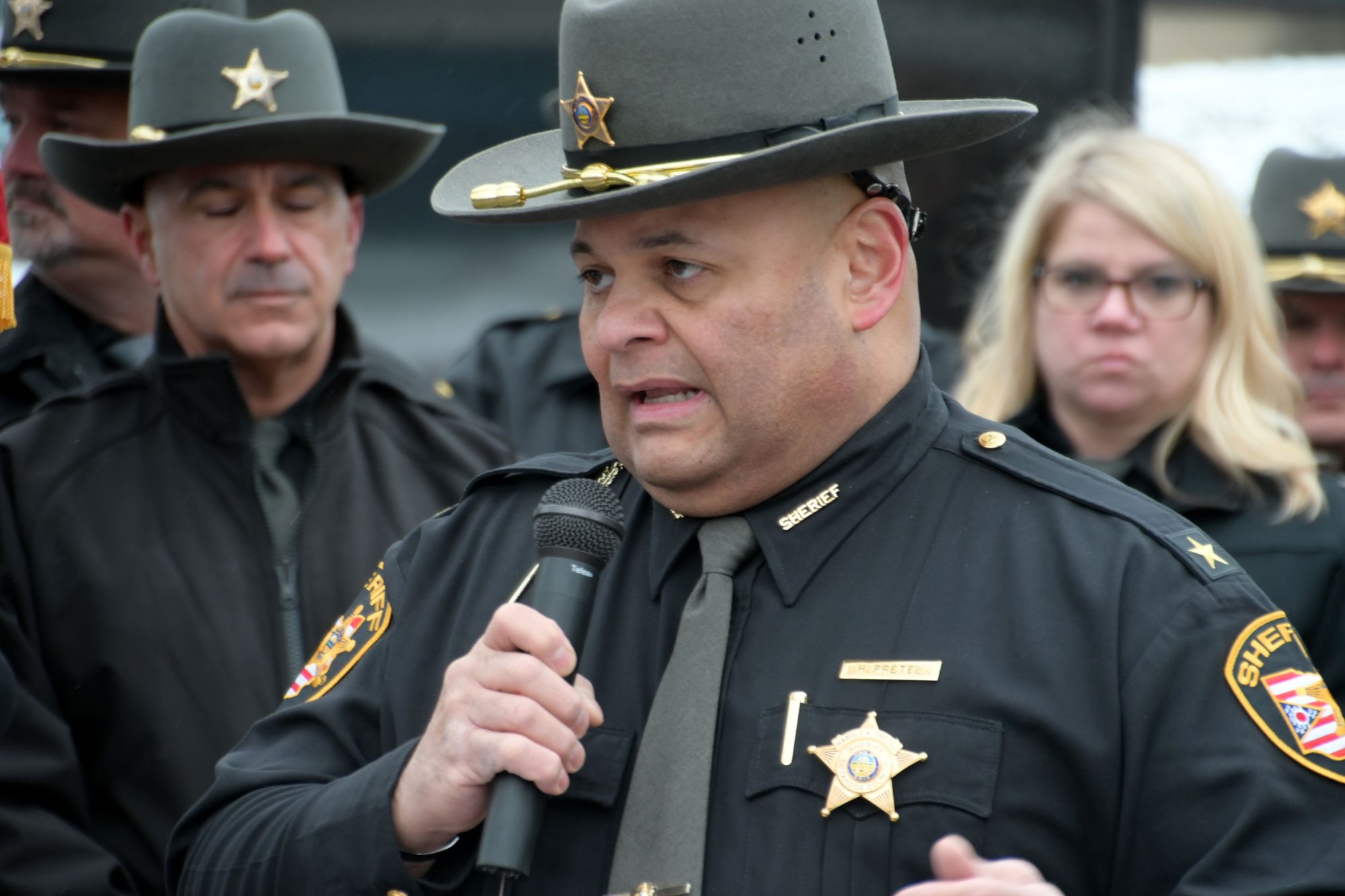 A man with medium-toned skin, wearing a star on his uniform, speaks into a microphone outdoors.