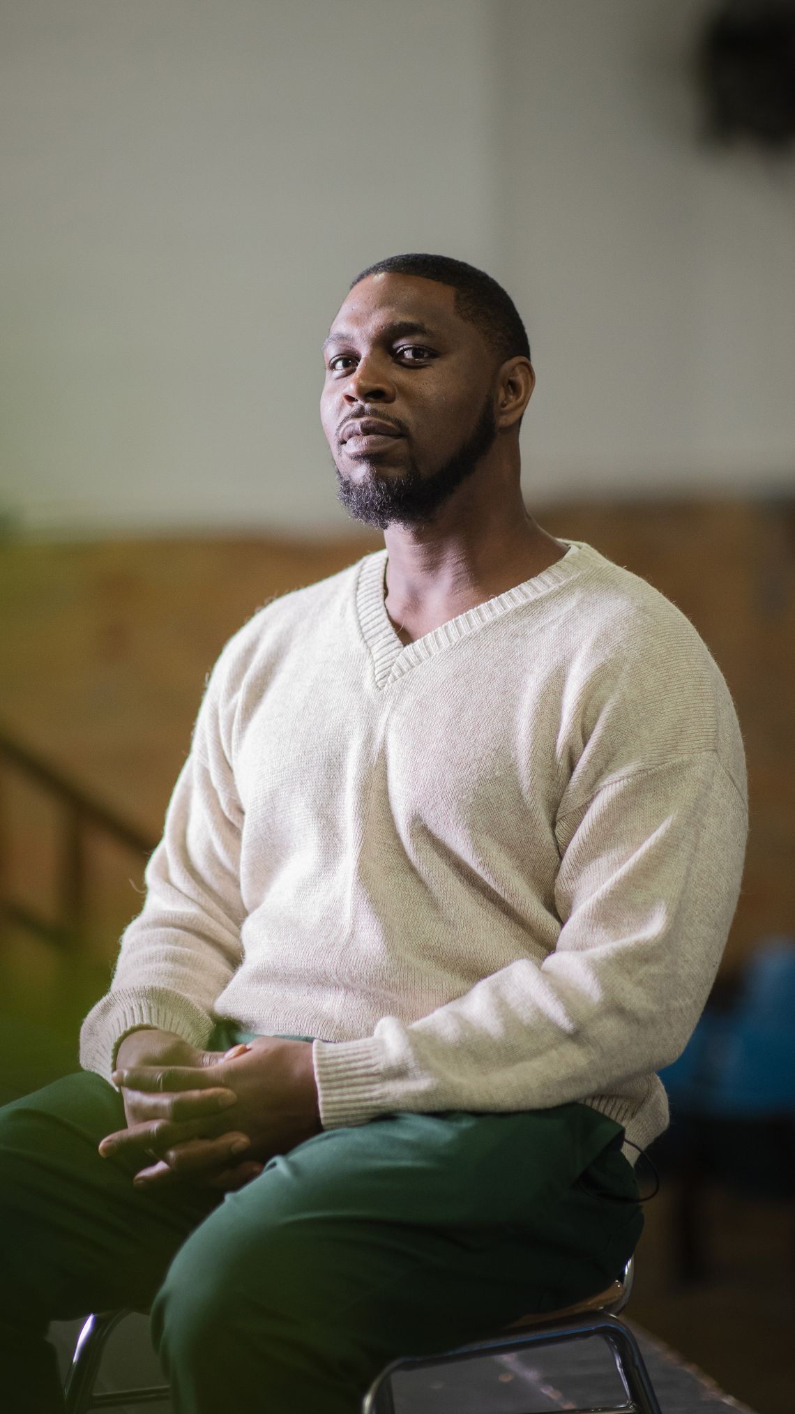 A Black man with a beard and wearing a beige sweater and green prison pants sits in a chair, staring at the camera with a smile on his face. 