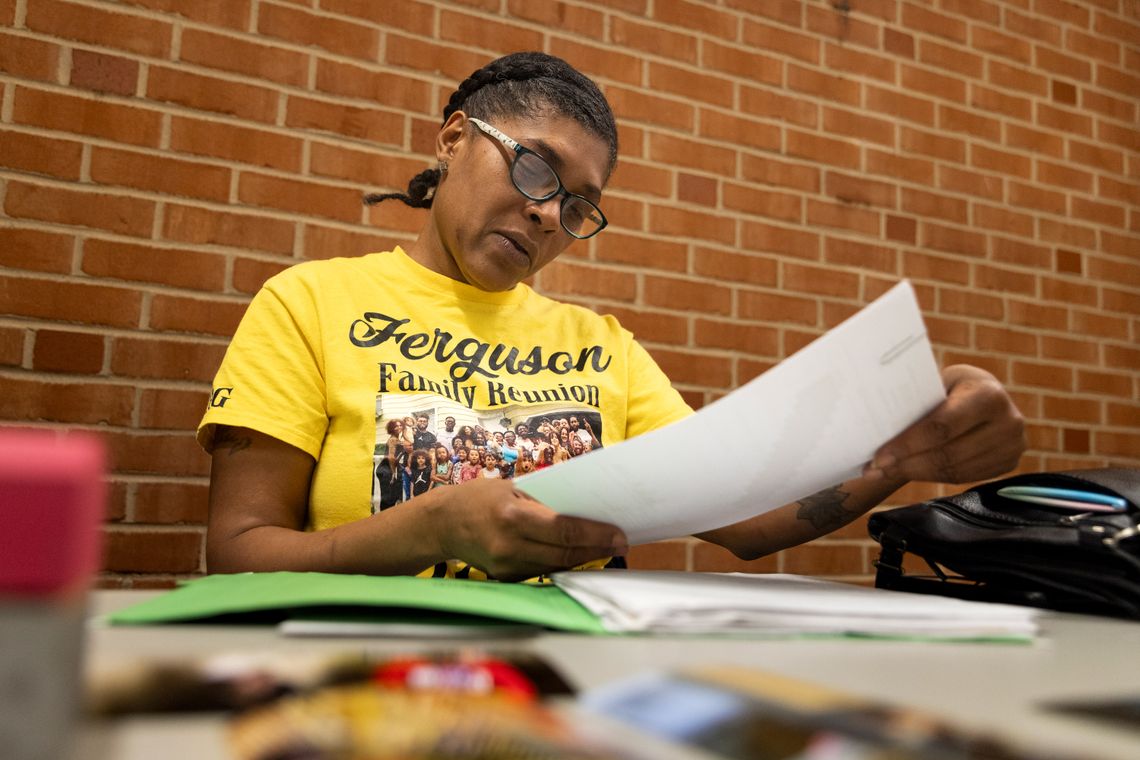 A photo of a middle-aged Black woman with glasses and her hair up looking at a piece of paper while sitting at a table in front of a brick wall. She is wearing a yellow shirt with a family photo under the words “Ferguson Family Reunion.”