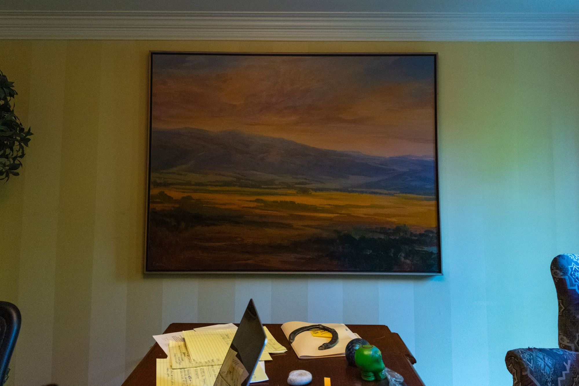 A painting showing a mountainside, orange clouds and blue sky hangs on a wall as light comes in from the right side. A table with a laptop and some papers is visible at the bottom of the photo.