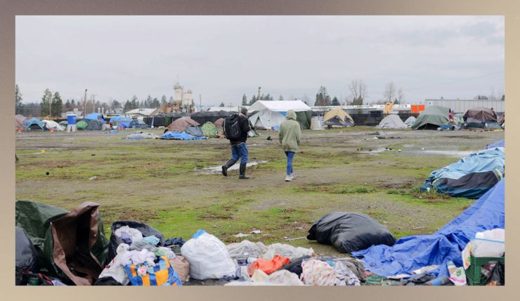 Two people walk through an encampment for unhoused people, which is on a field with some patches of grass and dirt.  Tents are visible both in the distance and in the foreground, and they stand beside the residents' belongings.    