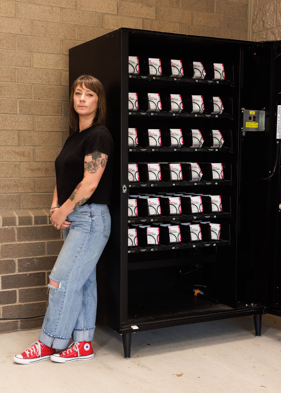 A photo of a White woman with brown hair, wearing a black t-shirt, blue jeans and red Converse sneakers. The woman stands against a black vending machine with white and pink Narcan boxes.