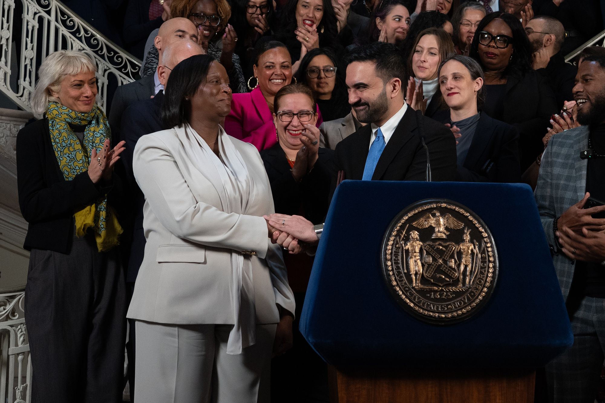 A photo shows a a Black woman wearing a light gray suit shaking the hands of an American man of South Asian descent wearing a black suit and blue tie.  They are standing behind a podium, and a group of people are standing behind them clapping.  