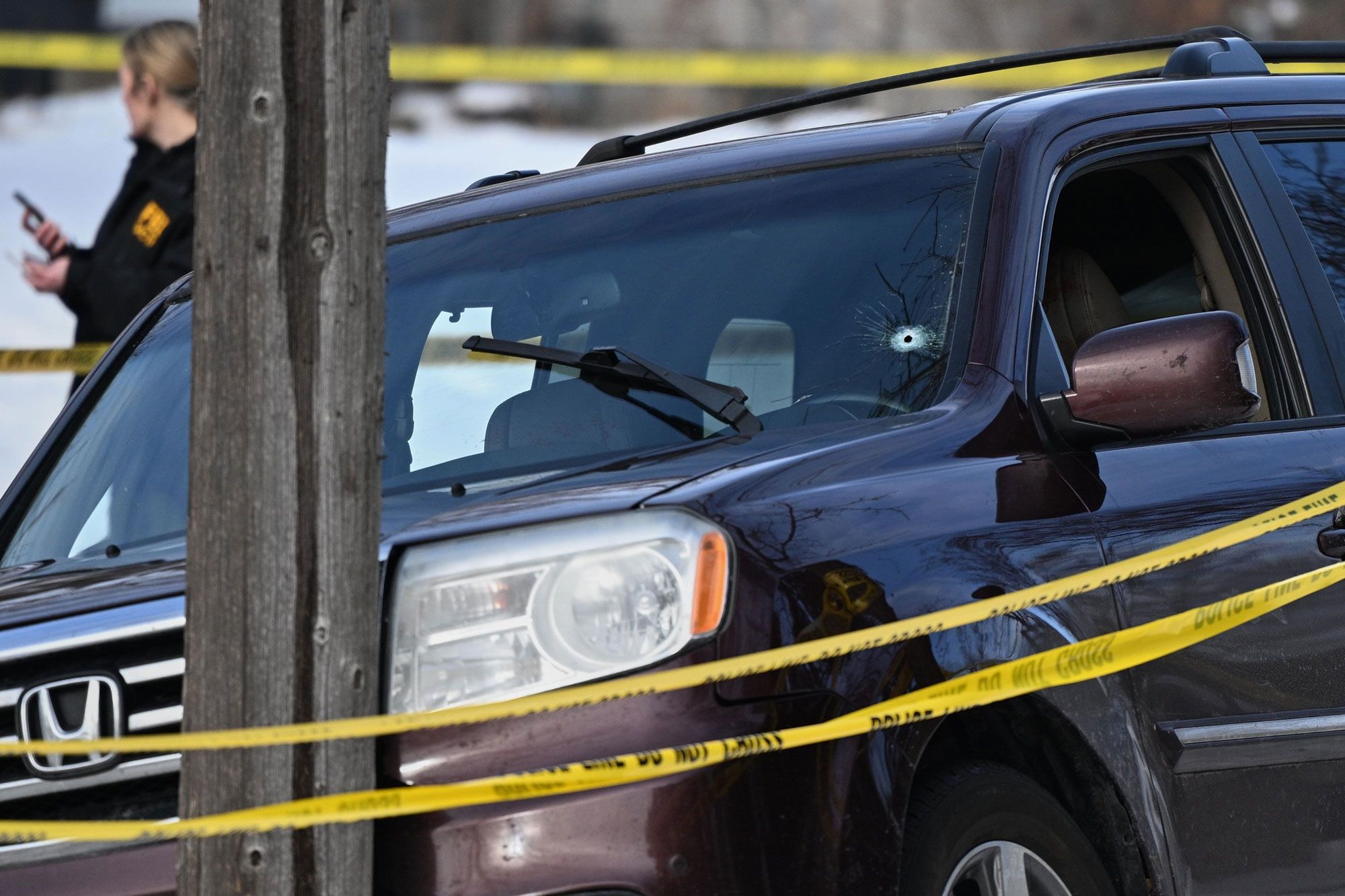 A bullet hole appears in the bottom left of the windshield of a dark-colored SUV that has struck a pole. In the foreground are two ribbons of police tape and in the background stands a person with a phone. 