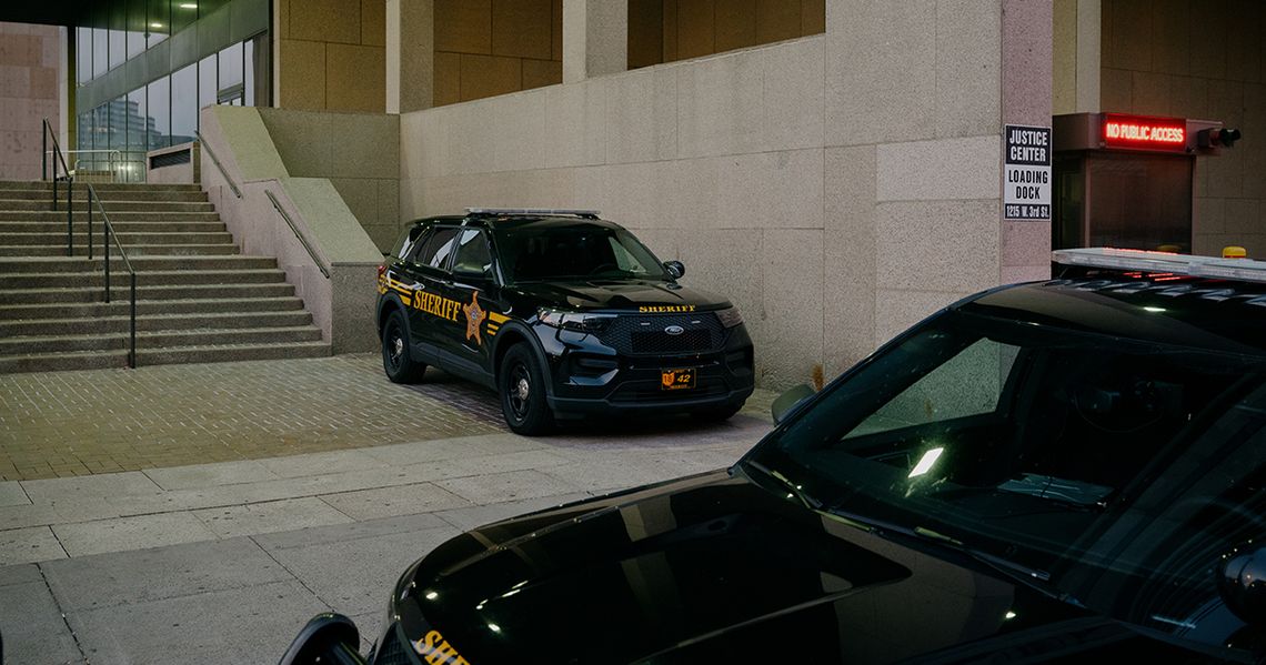 An empty black sheriff's vehicle, with the word "Sheriff" printed on the side in yellow paint, is parked near a beige-colored building. Another sheriff's vehicle is partly visible in the foreground. 