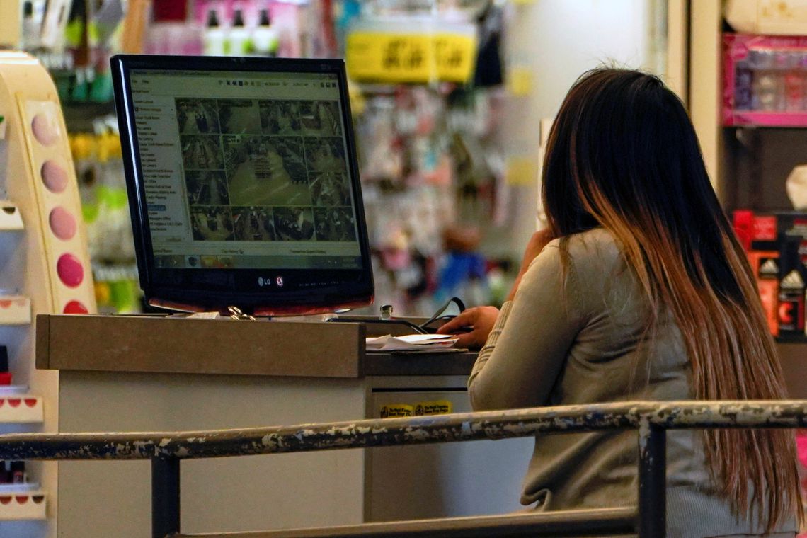 A woman with long dark hair with a gradient color and wearing a light gray long-sleeved shirt looks at security camera footage at a desk inside a store.