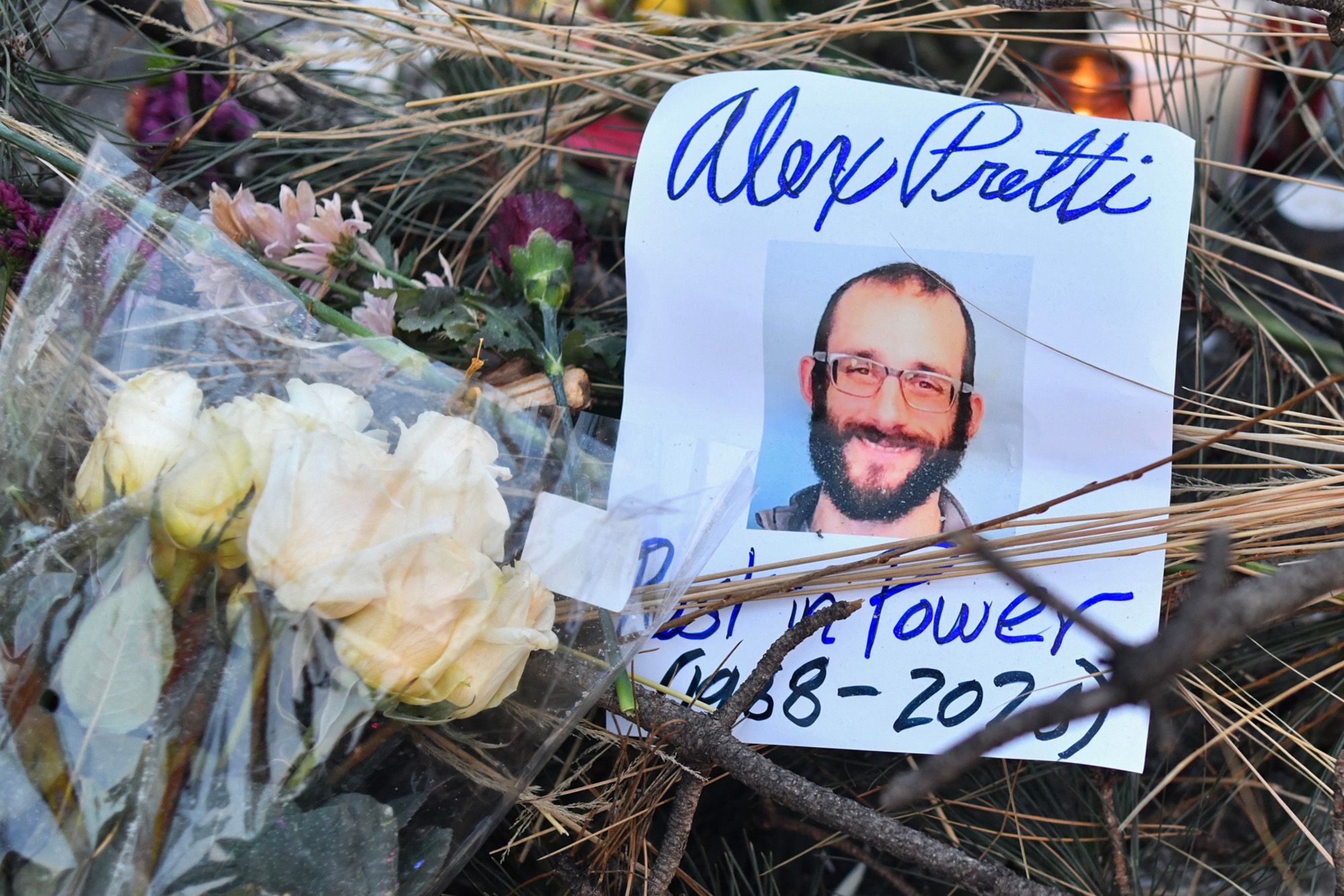 A photo shows a flyer with a head shot of a White man with a beard and glasses smiling and text that reads, "Alex Pretti, Rest in Power (1988-2026)." The flyer is placed in a memorial with flowers. 