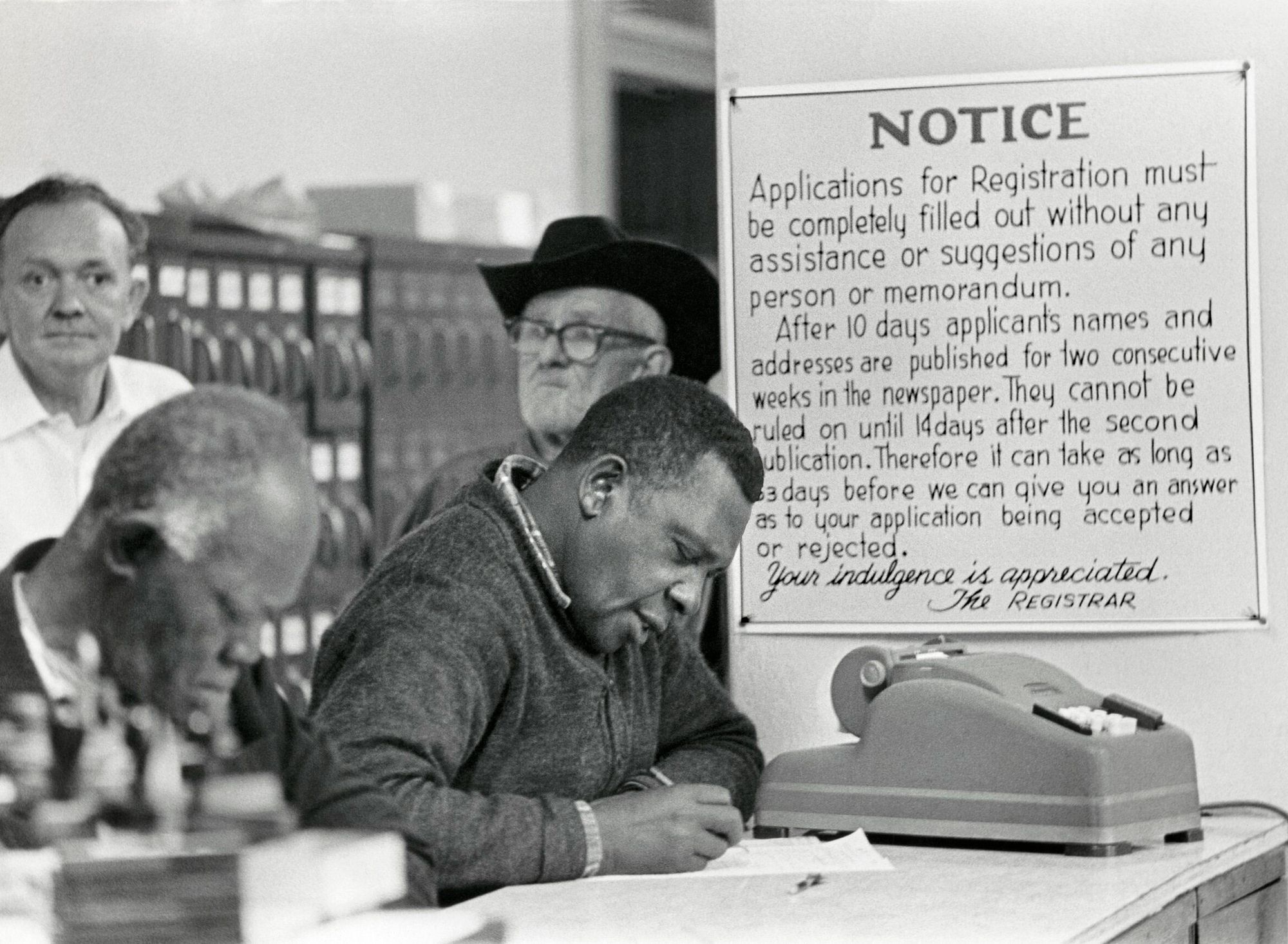 A black-and-white photo shows two Black men filling out forms at a desk, while two older White men stand in the background. Beside the men is a notice from the voting registrar. 