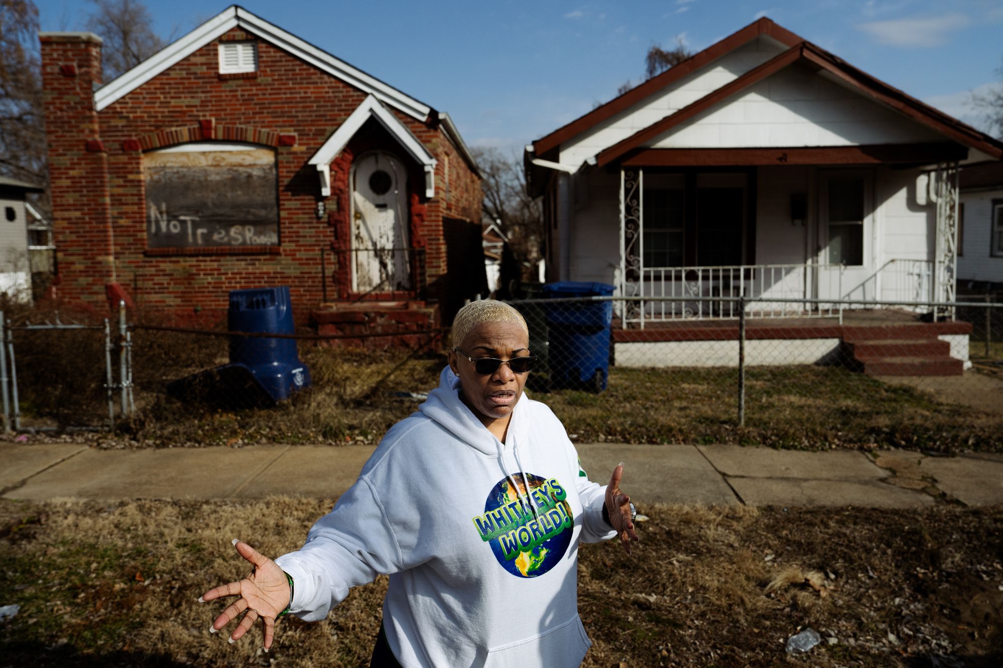 A Black woman with blonde hair gestures outside of an abandoned brick home and a home with white siding while wearing a white hoodie that says “Whitney’s World!” on it.