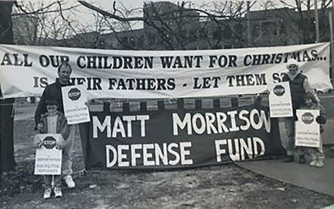 A black-and-white photo shows a man, a woman and two young children holding signs. They stand in front of two big signs that read, “All our children want for Christmas is their fathers — let them stay,” and “Matt Morrison defense fund.” 