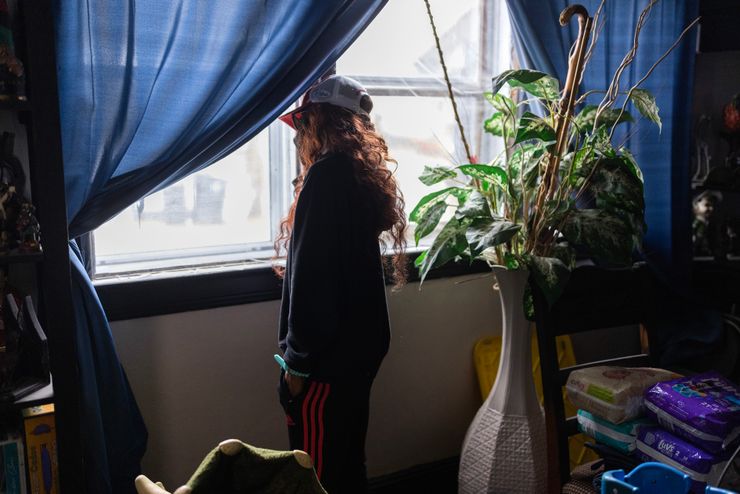 A woman with long, wavy red hair, whose face is not visible, looks out a window with blue curtains. Behind her is a tall vase with green leaves in it. A stack of diapers is on a chair.  