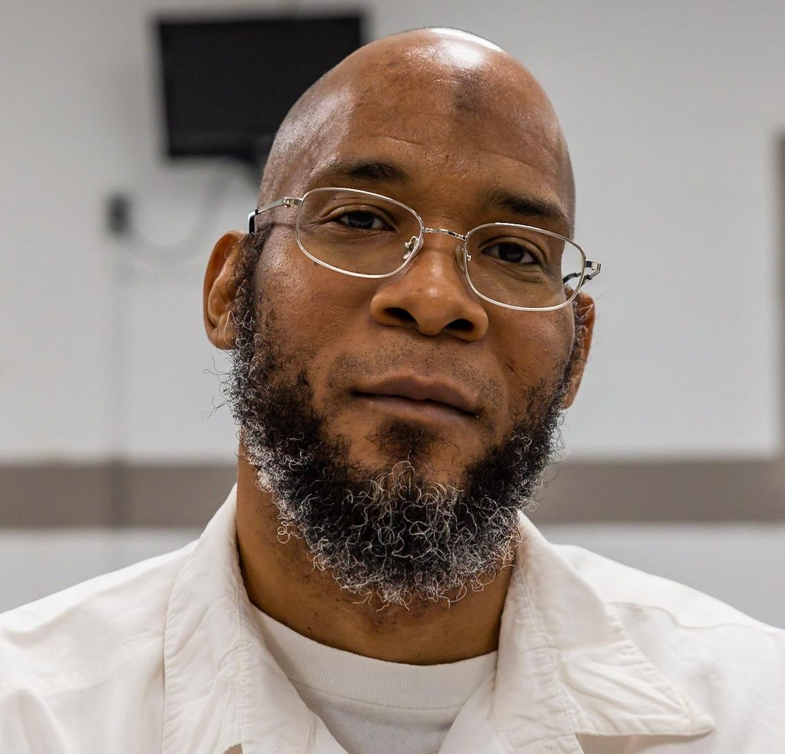 A photo shows a Black man with a beard, wearing glasses and a white prison uniform, posing for a portrait.