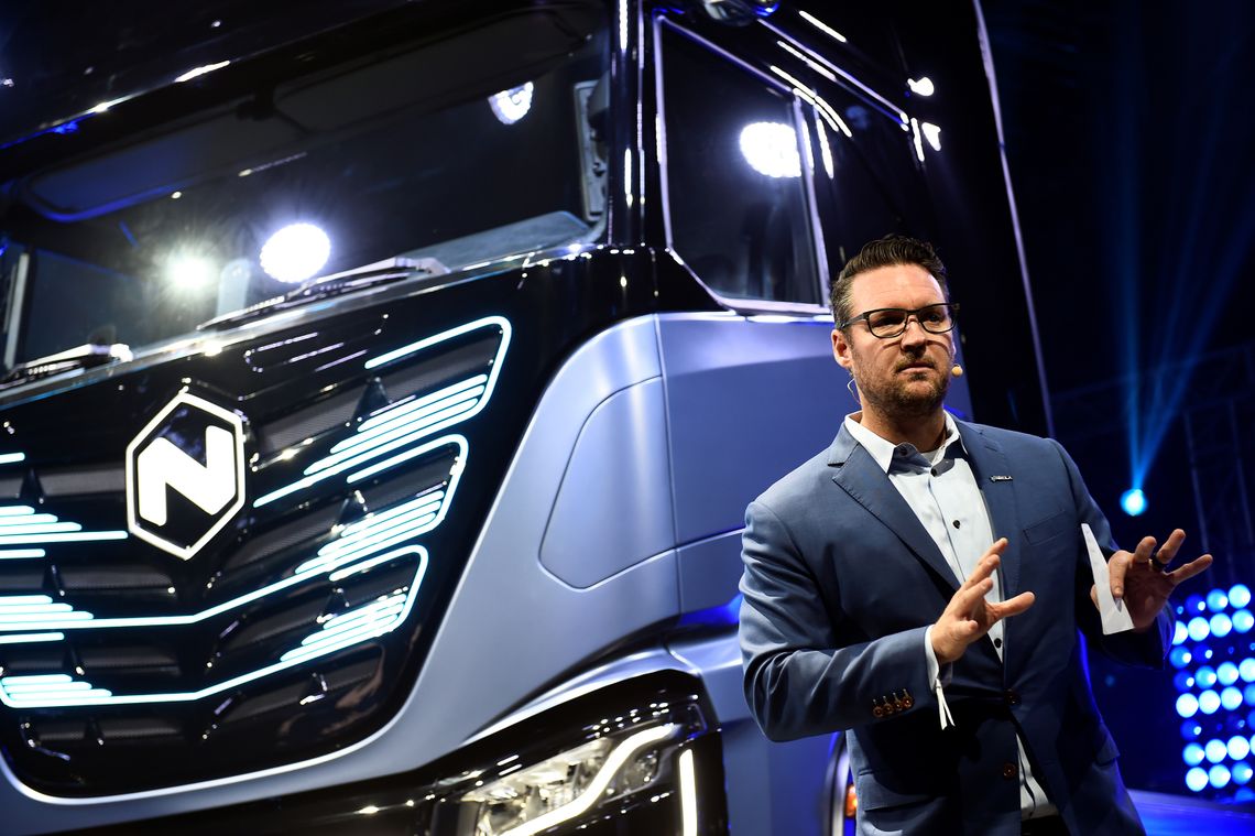 Trevor Milton, a man with light-toned skin, wearing glasses and a suit, stands indoors in front of a truck with blue and black panels.