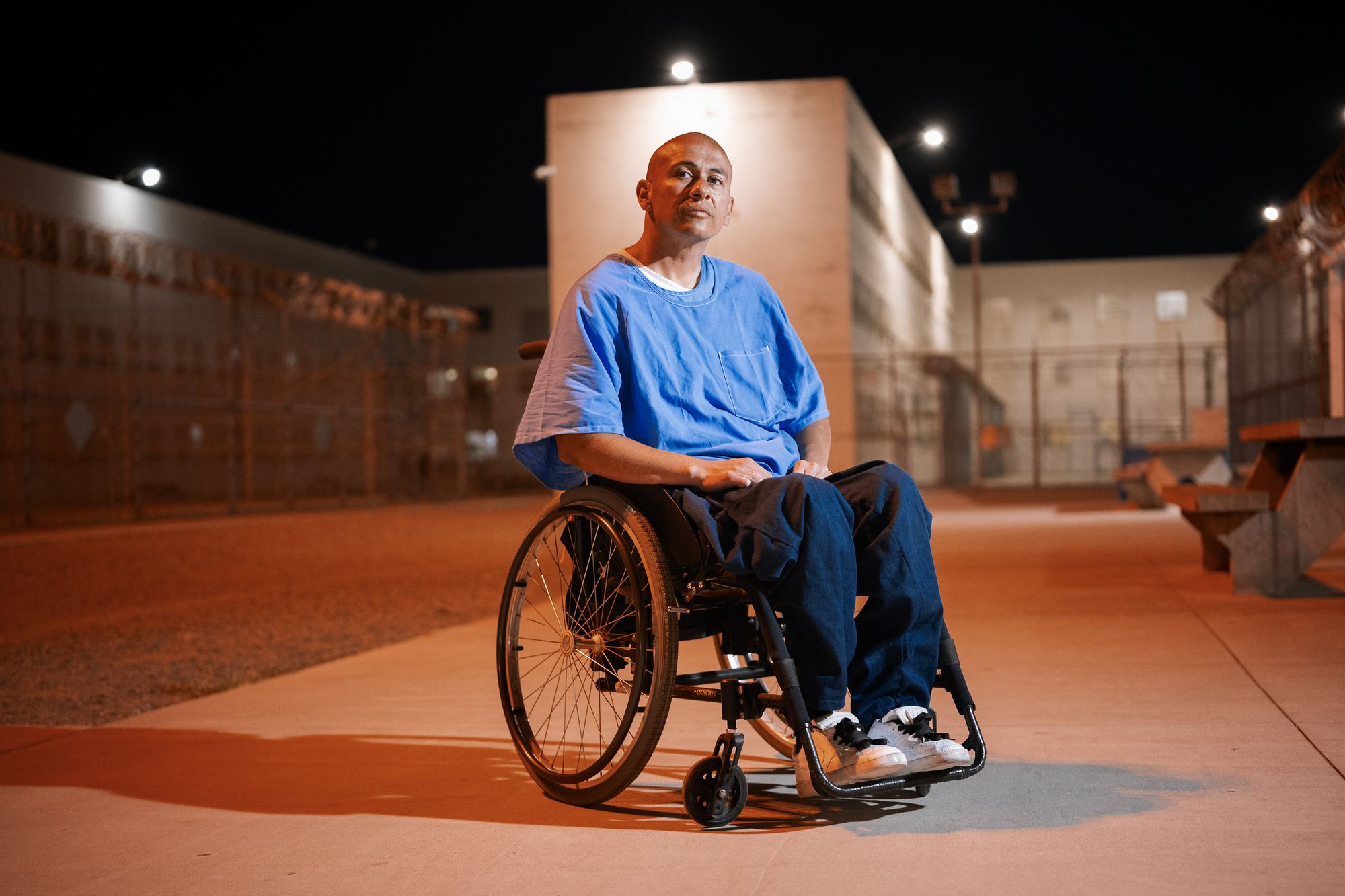 A photo shows a man with medium-toned skin, wearing a blue shirt and dark blue pants, sitting outdoors in a wheelchair. It is dark outside, and part of the prison yard is visible. 