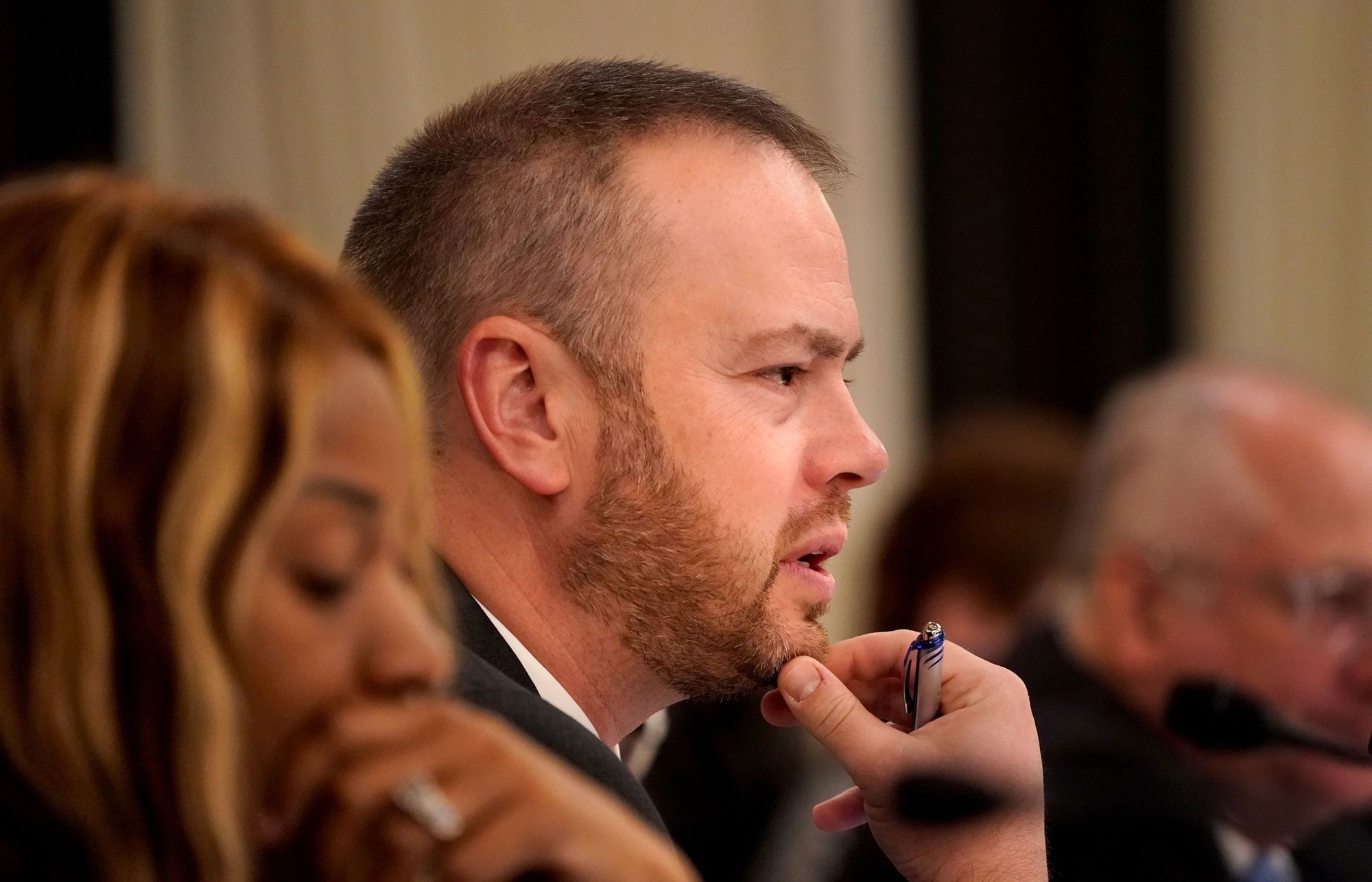 A White man holds his chin while listening to someone off-camera.