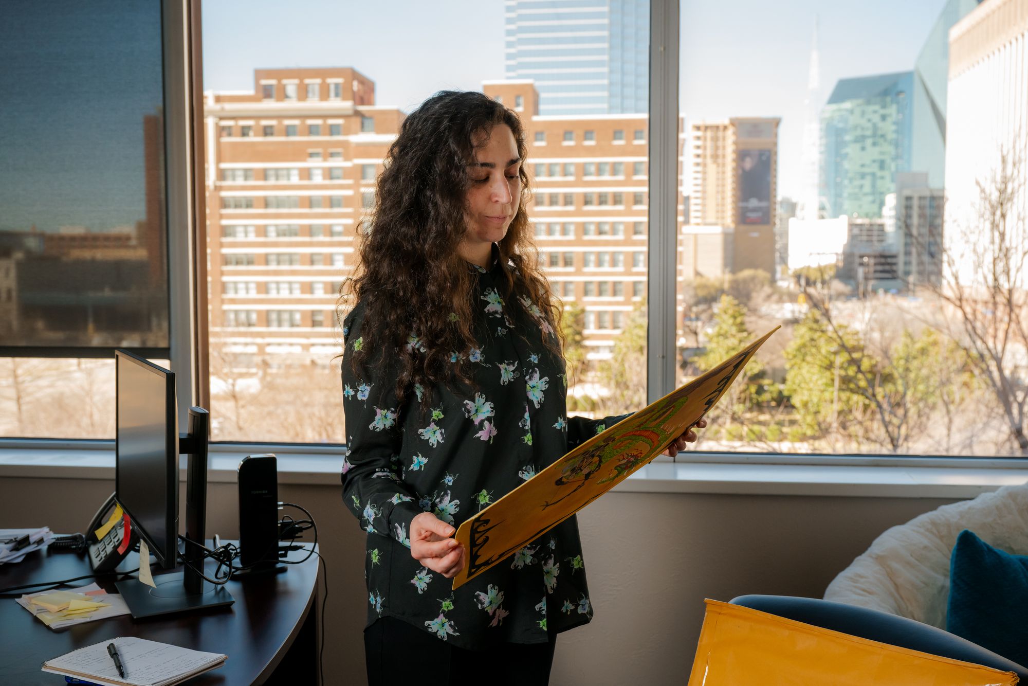 A photo shows a White woman with long curly hair wearing a floral long-sleeved shirt, standing in front of windows, holding a drawing. To the left is a desk with a computer monitor, and to the right is a sofa. 