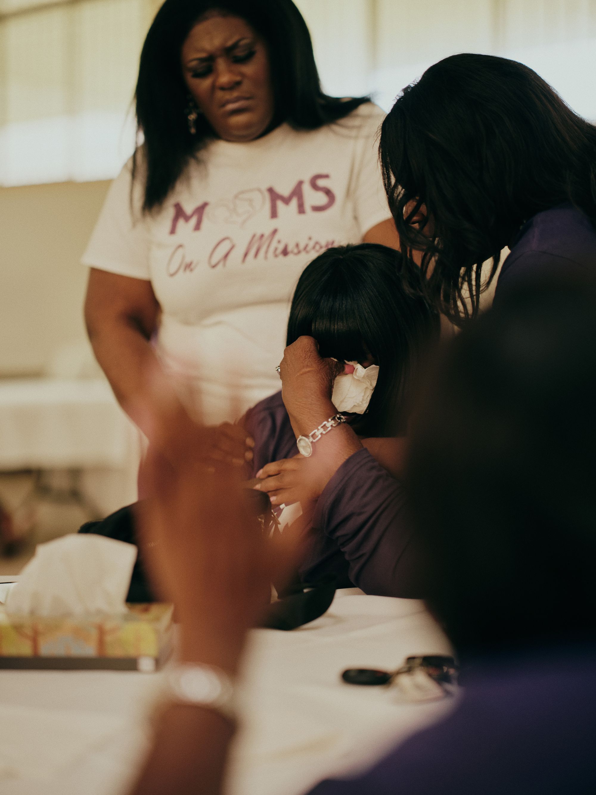 Shirley Pollard, a Black woman, holds a tissue to her face as one woman holds her, and another woman stands behind her.