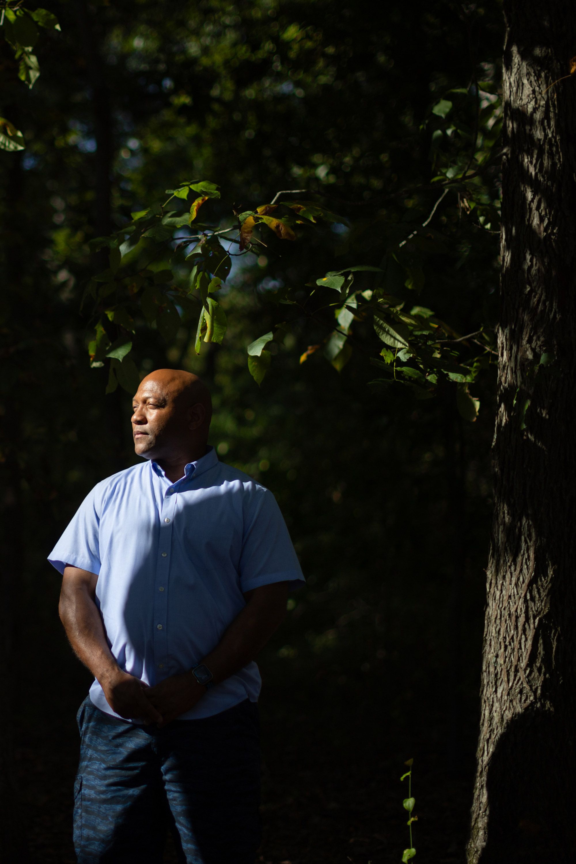 A photo of a bald Black man in a blue button-down shirt and blue shorts. The man is looking to the side toward the sun while standing next to a tree.