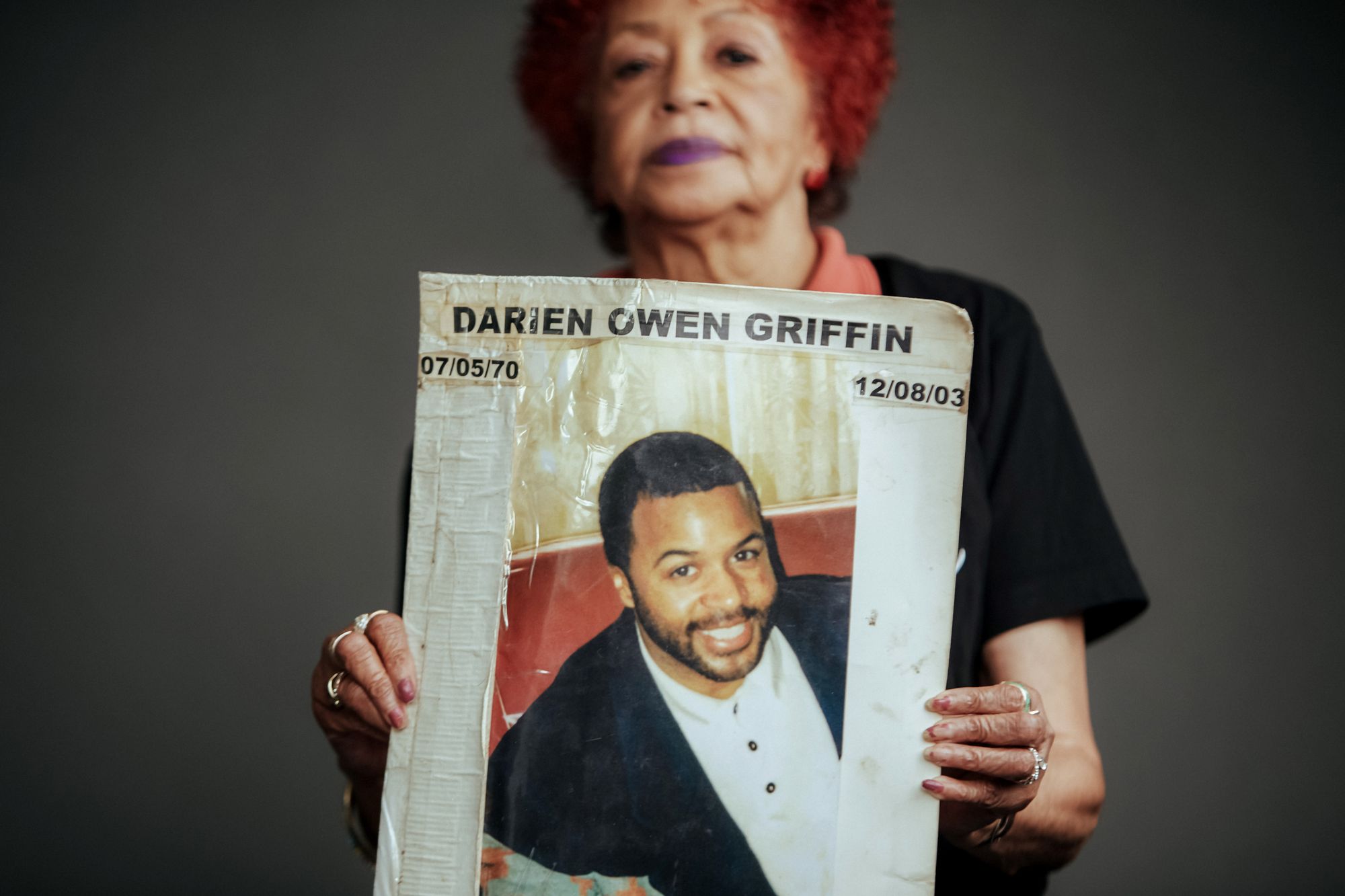 Dr. Patricia Griffin, a woman with light brown skin, holds out in front of a poster of her son Darien Owen Griffin, who was killed in 2003. On the poster, it states Darien's name and his dates of birth and death. 