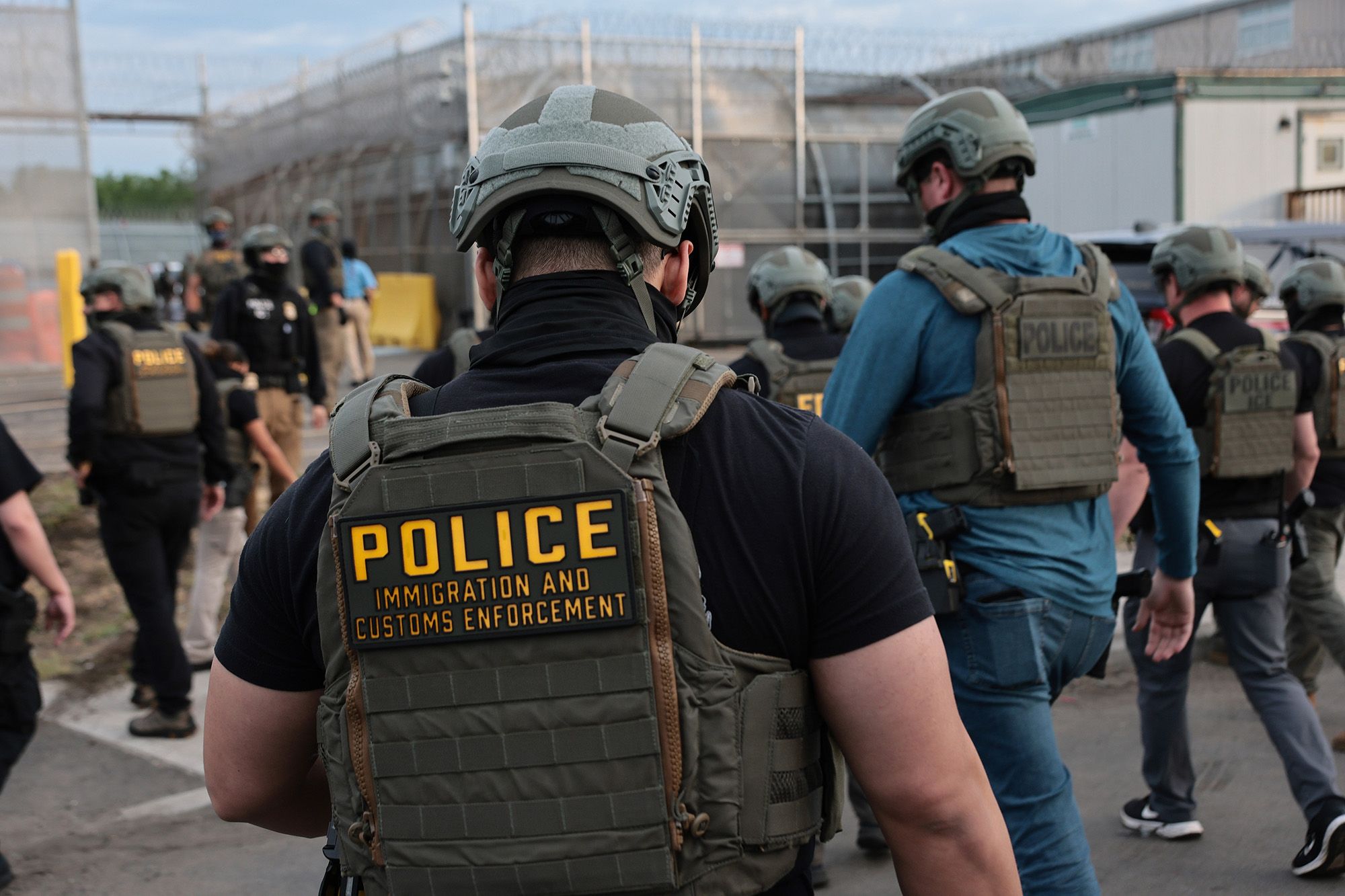 A photo shows several Immigration and Customs Enforcement agents, wearing helmets, masks and vests, standing in front of a gray building surrounded by barbed wire fences. 