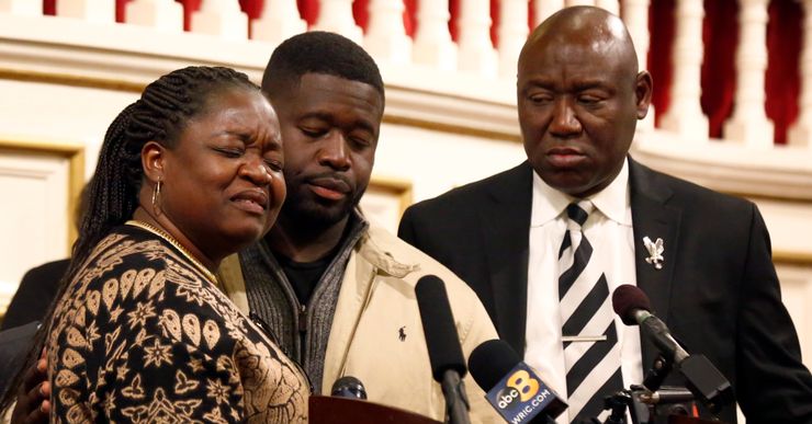 Caroline Ouko, a Black woman, and Leon Ochieng, and Ben Crump, two Black men, stand before a podium.  Ouko and Ochieng have their eyes closed and have emotional expressions on their faces.  