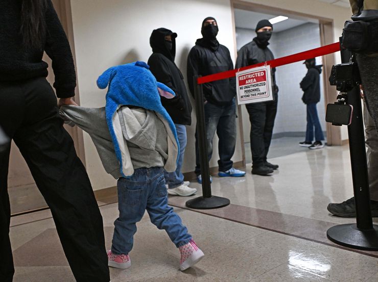 A photo shows a woman holding her child's hand as she walks down a hallway. On the right, ICE masked agents are standing against the wall.