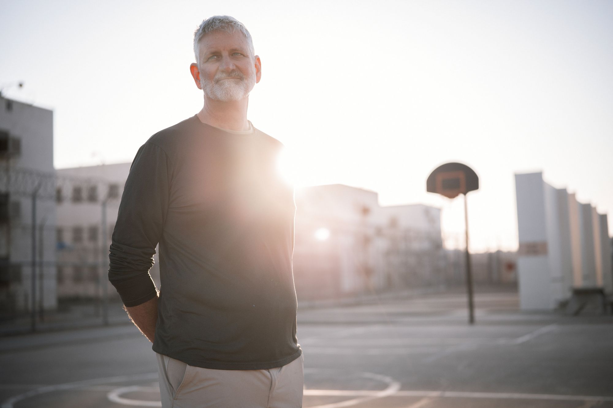 A photo shows a White man with a gray beard and hair, wearing a long-sleeved shirt, standing on the outdoor basketball court in the morning. 