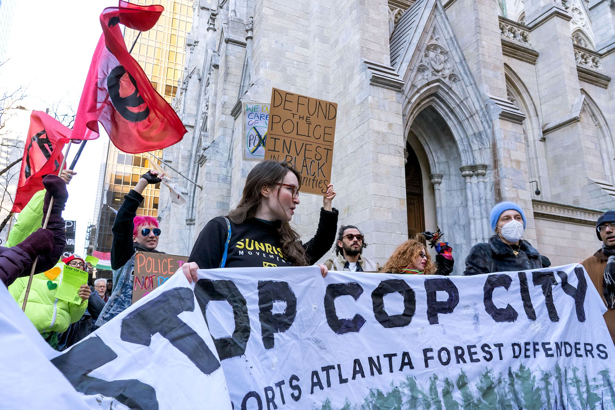 Several protesters hold a banner that reads "Stop Cop City," as they stand in front of a church. One protester in the background holds a sign that reads, "Defund the police. Invest in Black communities."  