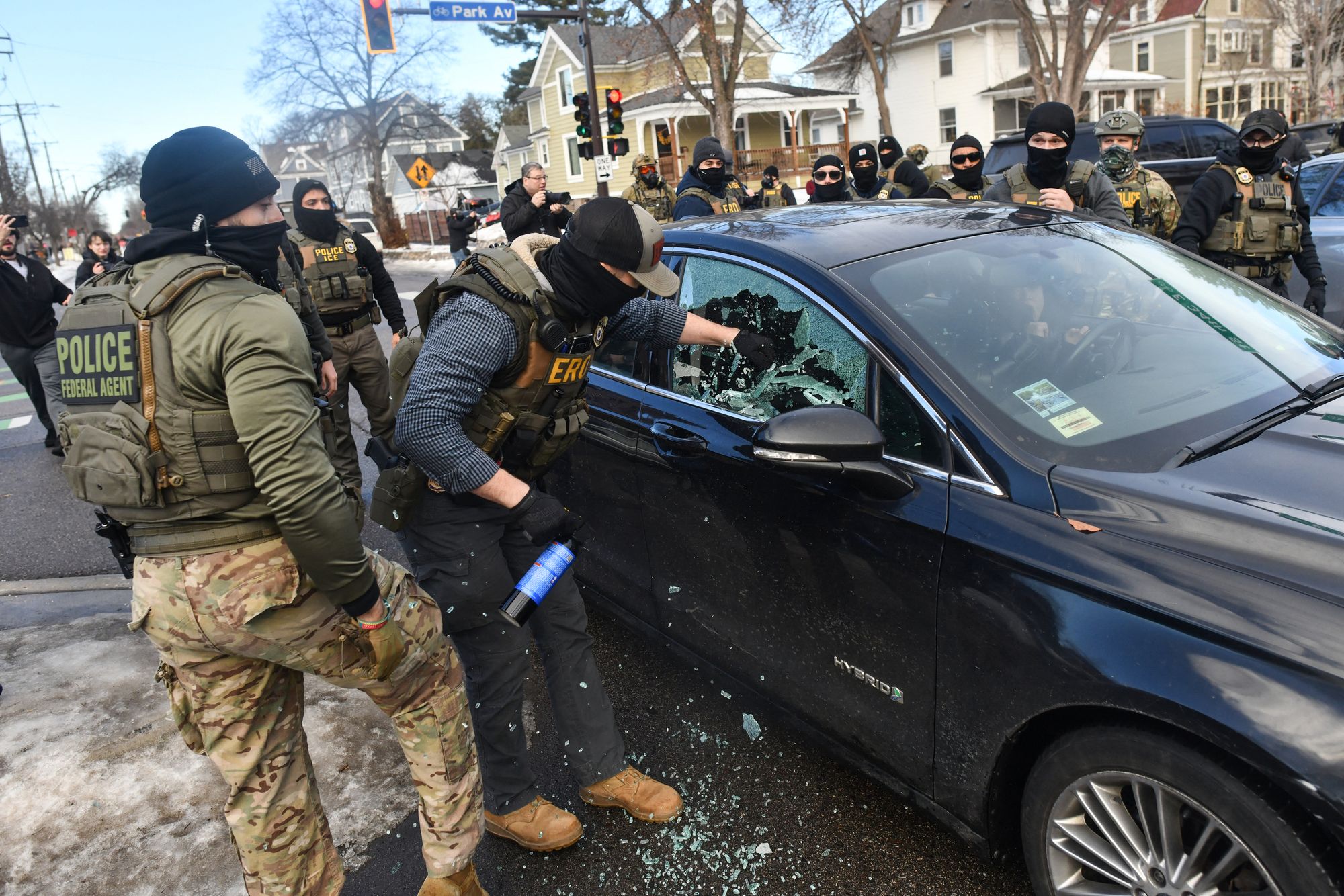 A photo shows a woman in the driver's seat of a car looking shocked as a federal agent smashes the passenger side window. A number of other federal agents stand by and watch. Some houses are visible in the back of the photo. 