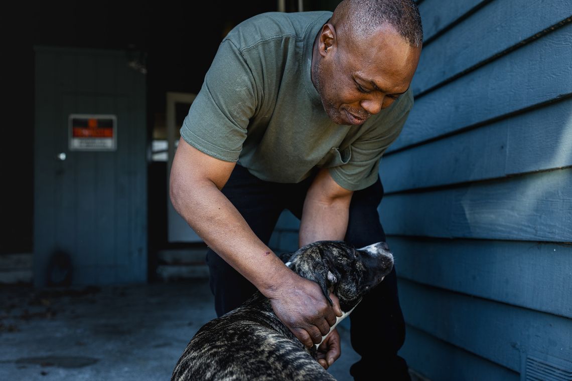 A Black man, wearing a jade green T-shirt, smiles as he pets his dog outside his home. 