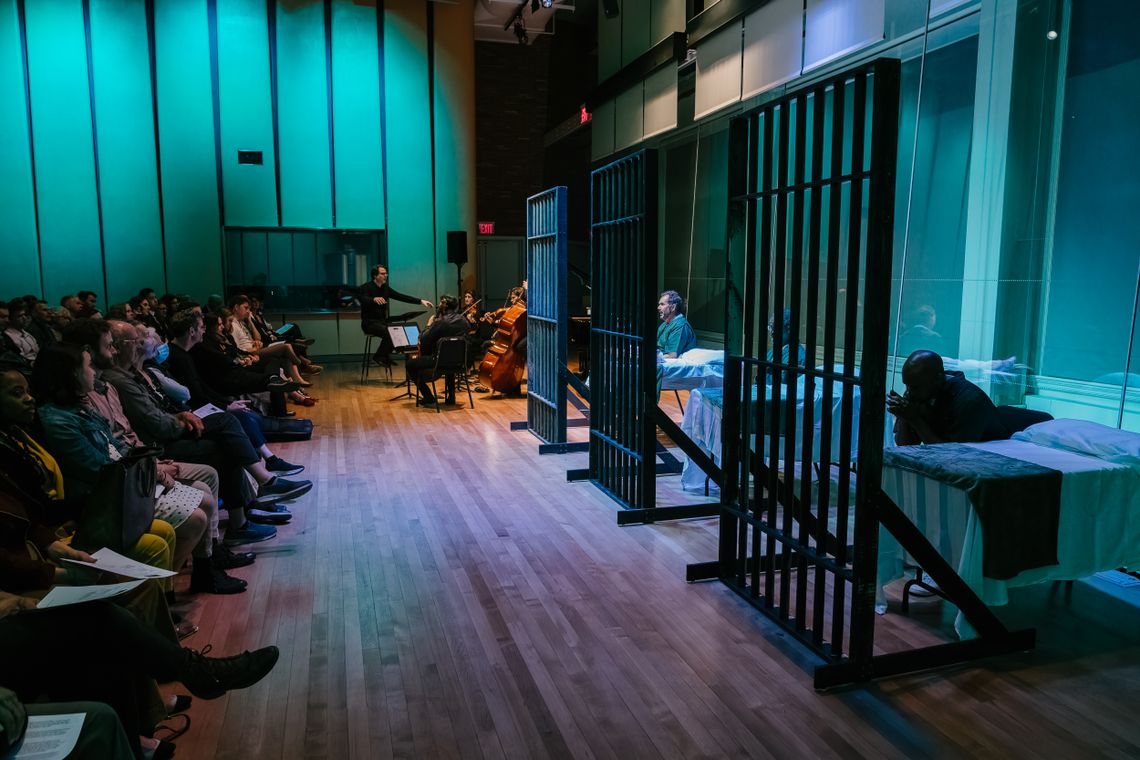 A photo shows a group of instrumentalists and three performers dressed in green prison uniform costumes, sitting or lying on beds, each behind a panel of prison bars. On the left are seated audience members.