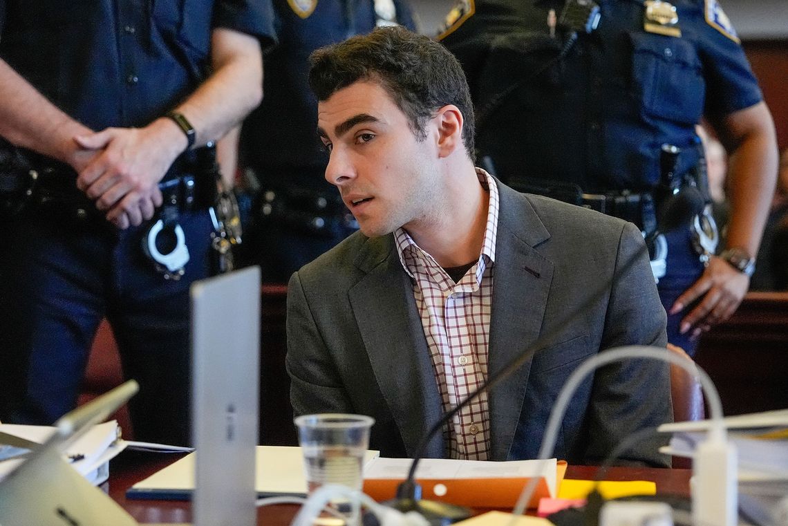 A photo shows Luigi Mangione sitting at a courtroom table, wearing a gray blazer and red-checkered shirt, leaning to his right and engaging in conversation with an attorney out of the frame. Behind him are two court officers dressed in navy blue uniforms, with a pair of shiny handcuffs attached to the officers’ waists.