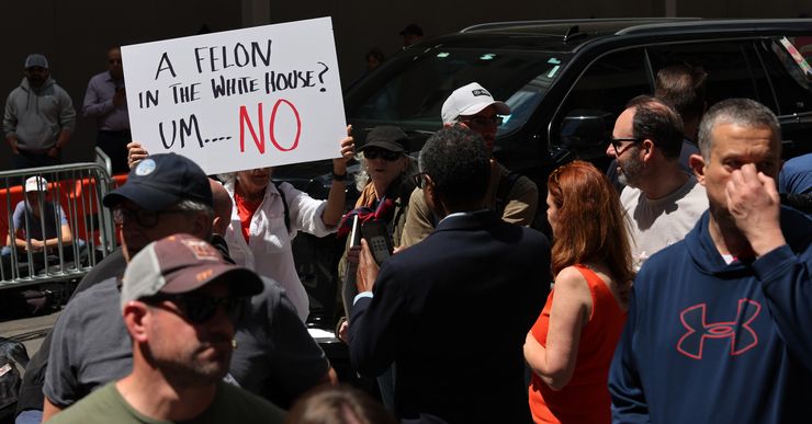 A group of people stand outside, with a vehicle nearby.  A person holds a sign that says: "A felon in the White House? Um...NO". 