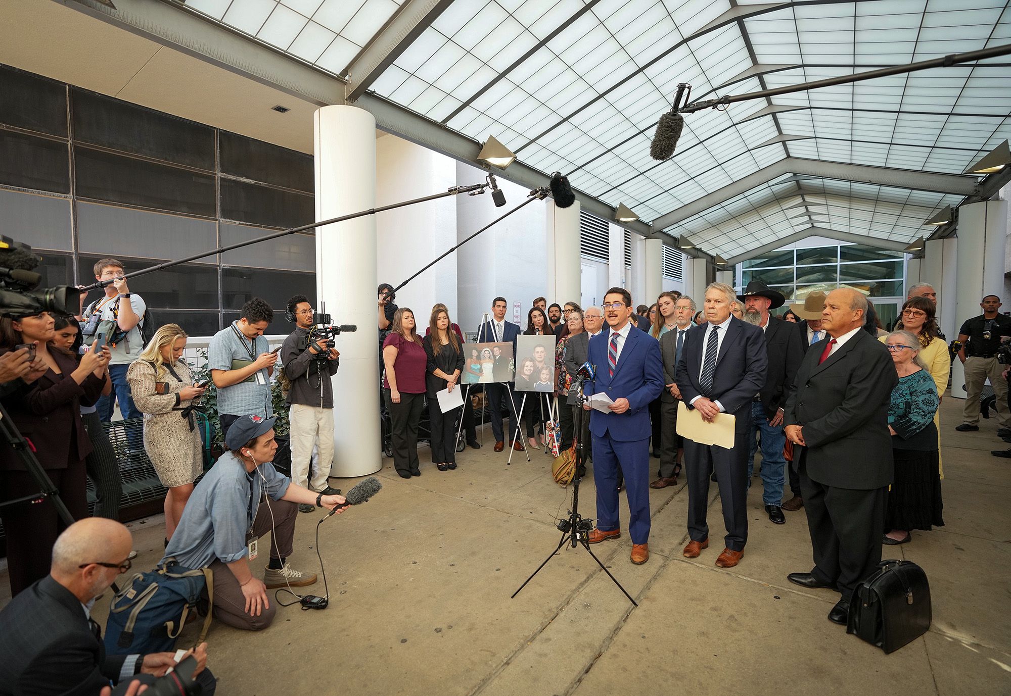 Travis County District Attorney José Garza, a Mexican man in a blue suit, speaks to reporters while a group of people stand behind him with portraits on printed boards.  
