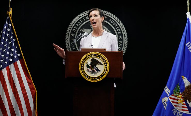 A White woman in a gray suit delivers a speech at a podium with the federal Bureau of Prisons logo on it. In the background, an American flag can be seen on the left and the U.S. Department of Justice flag on the right. 