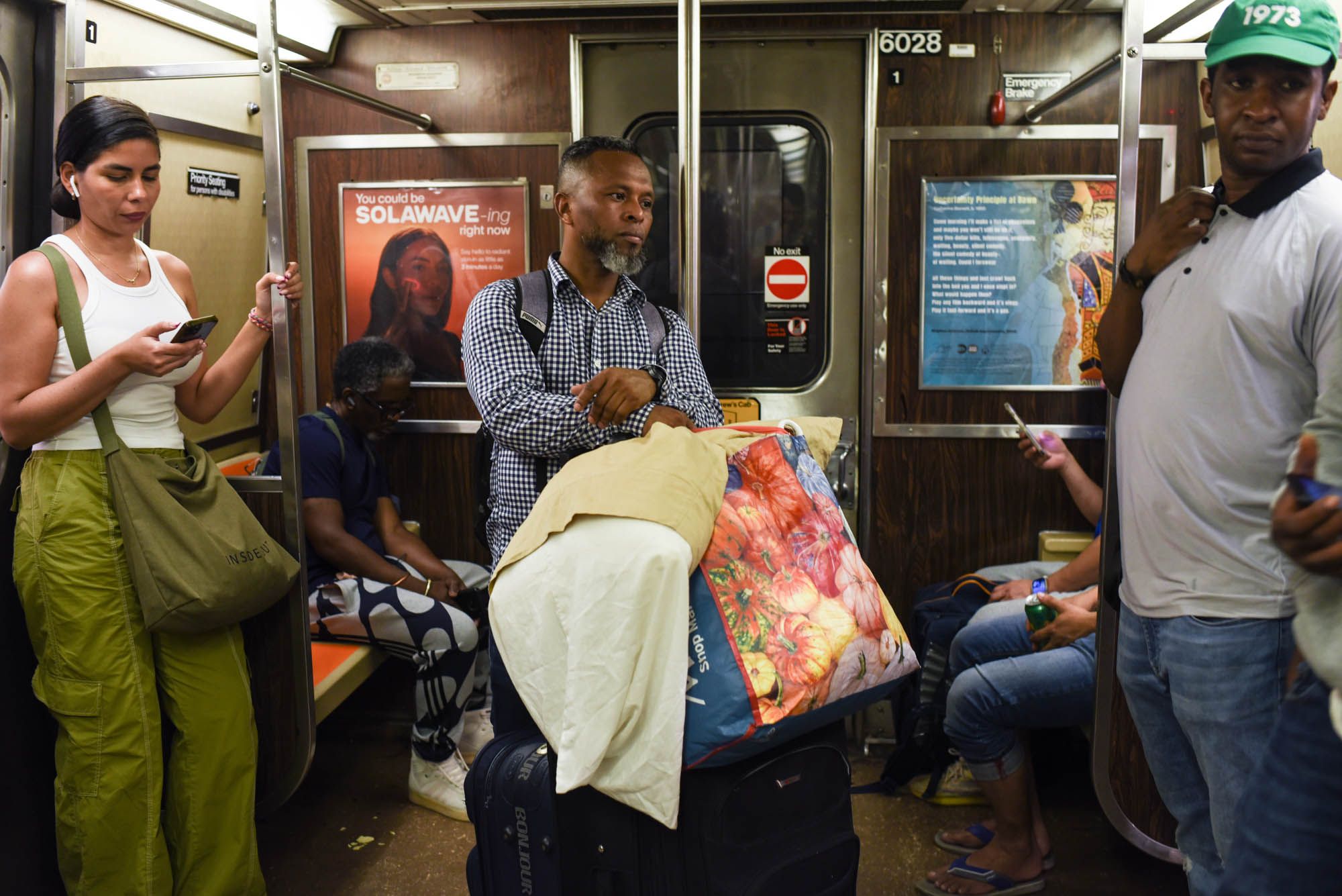 A medium dark-skinned Venezuelan man leans against a metal pole on a crowded subway, holding a large shopping bag and a pillow, as they rest atop a black, rolling suitcase. The man looks off to the side and downwards.