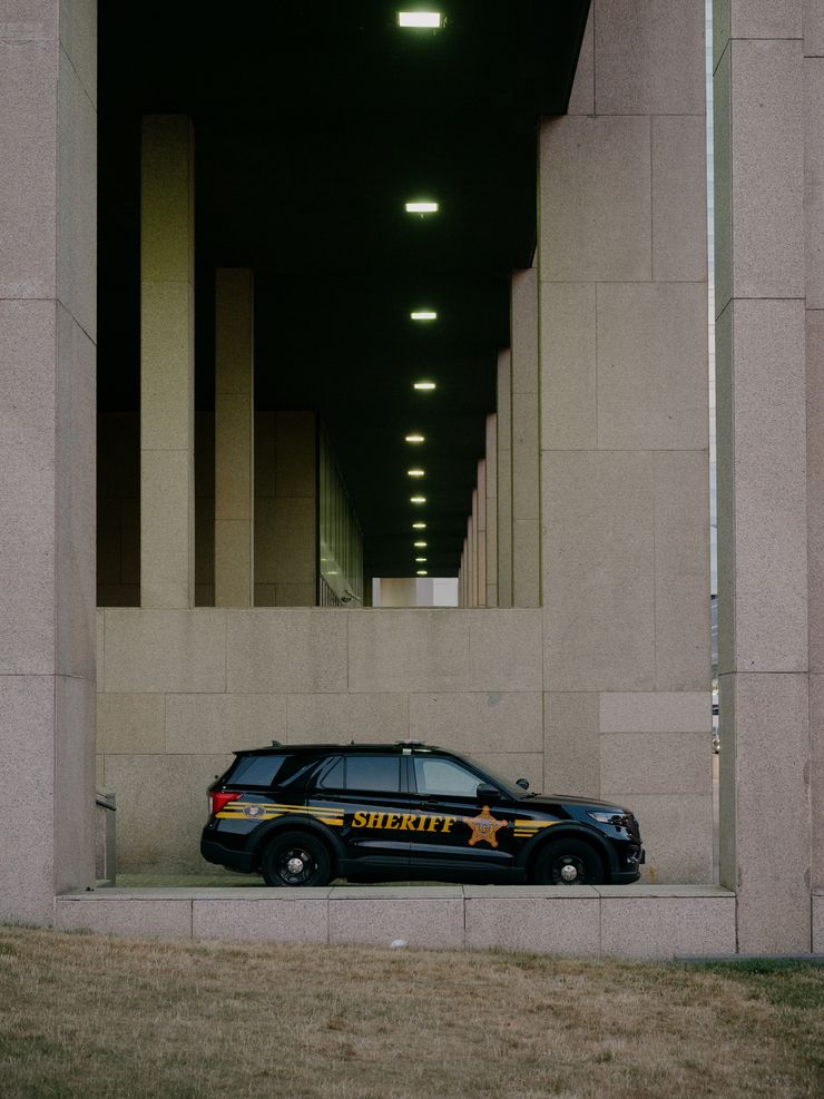 An empty black sheriff's vehicle, with the word "Sheriff" printed on the side in yellow paint, is parked at a beige-colored building with two columns on either side.  