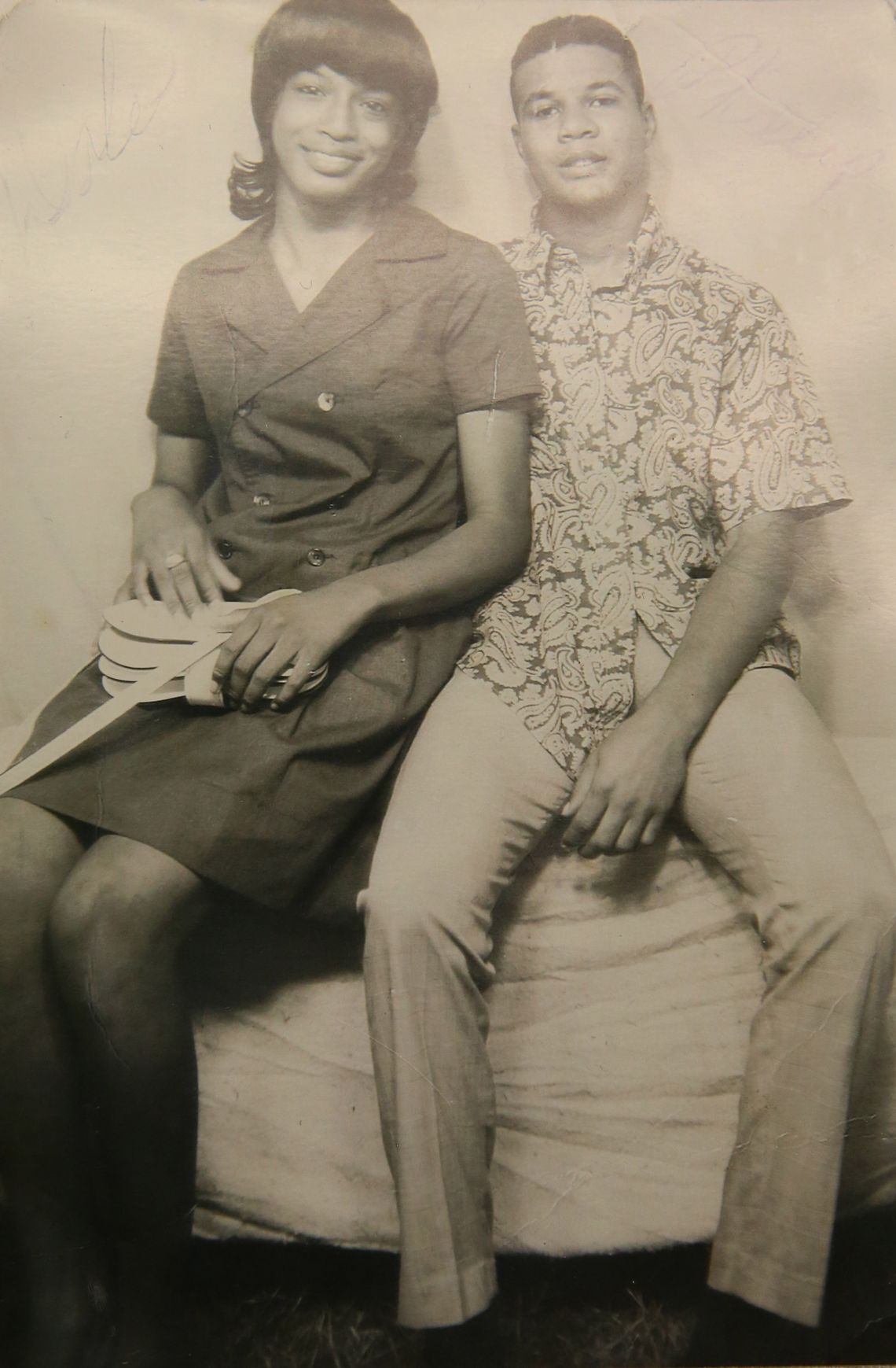 A sepia-toned photograph shows a couple — a Black woman in a dark knee-length dress, and a Black man in a patterned button-down shirt — sitting for a portrait. 