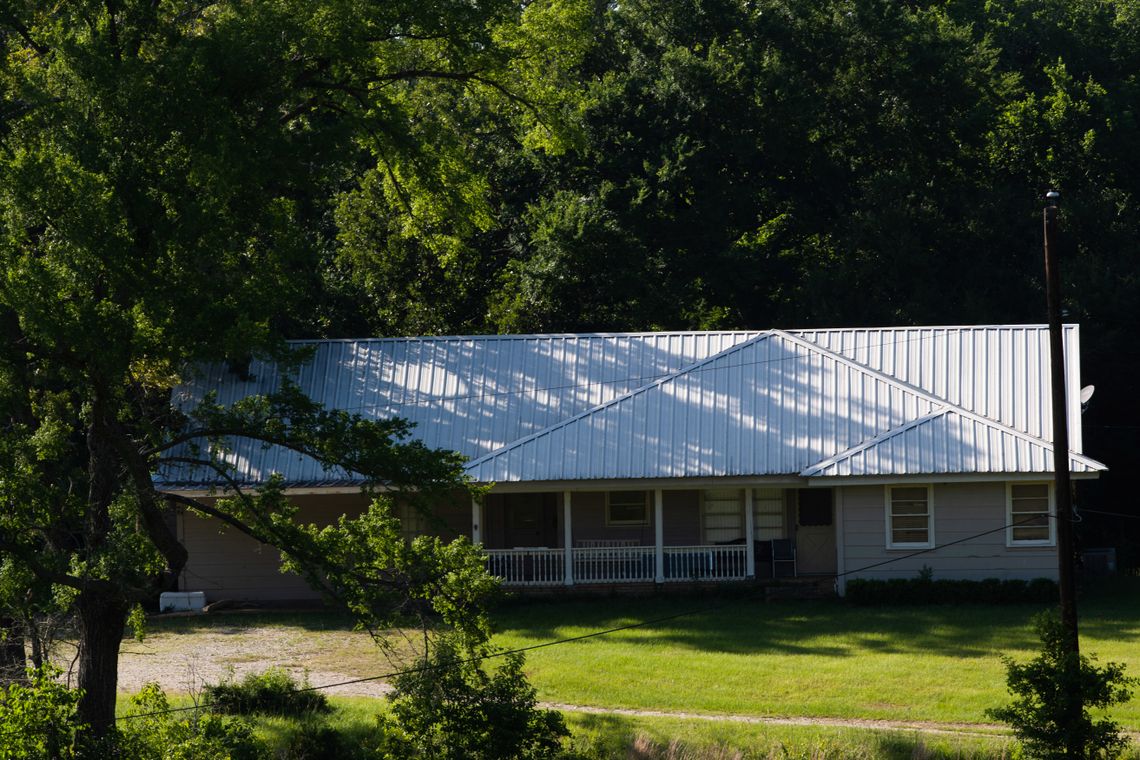 A one-story house sits behind a green lawn and among trees. 