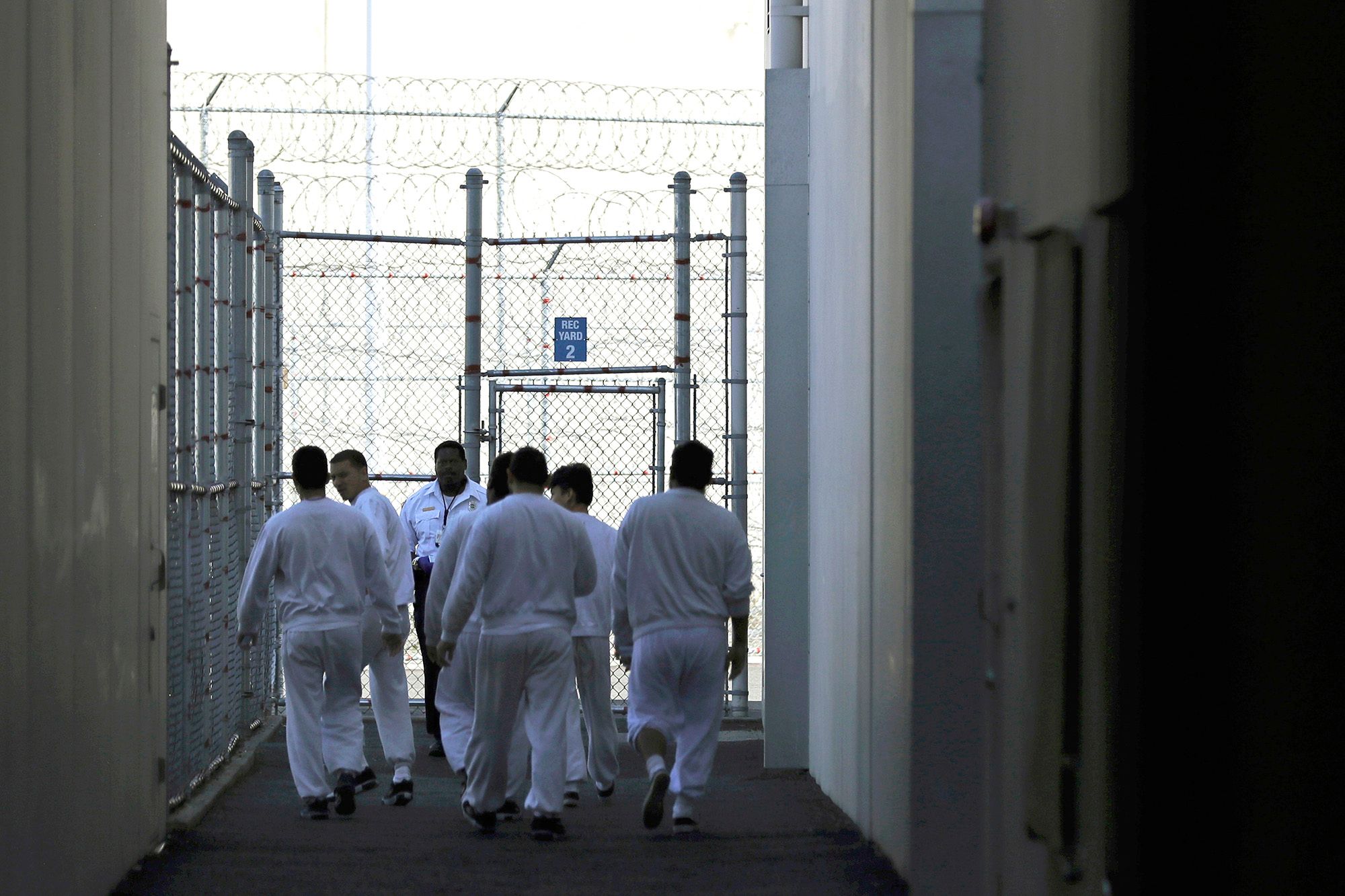 A group of men wearing white uniforms, seen from the back, walk toward a fenced area. A guard stands by the entrance. 